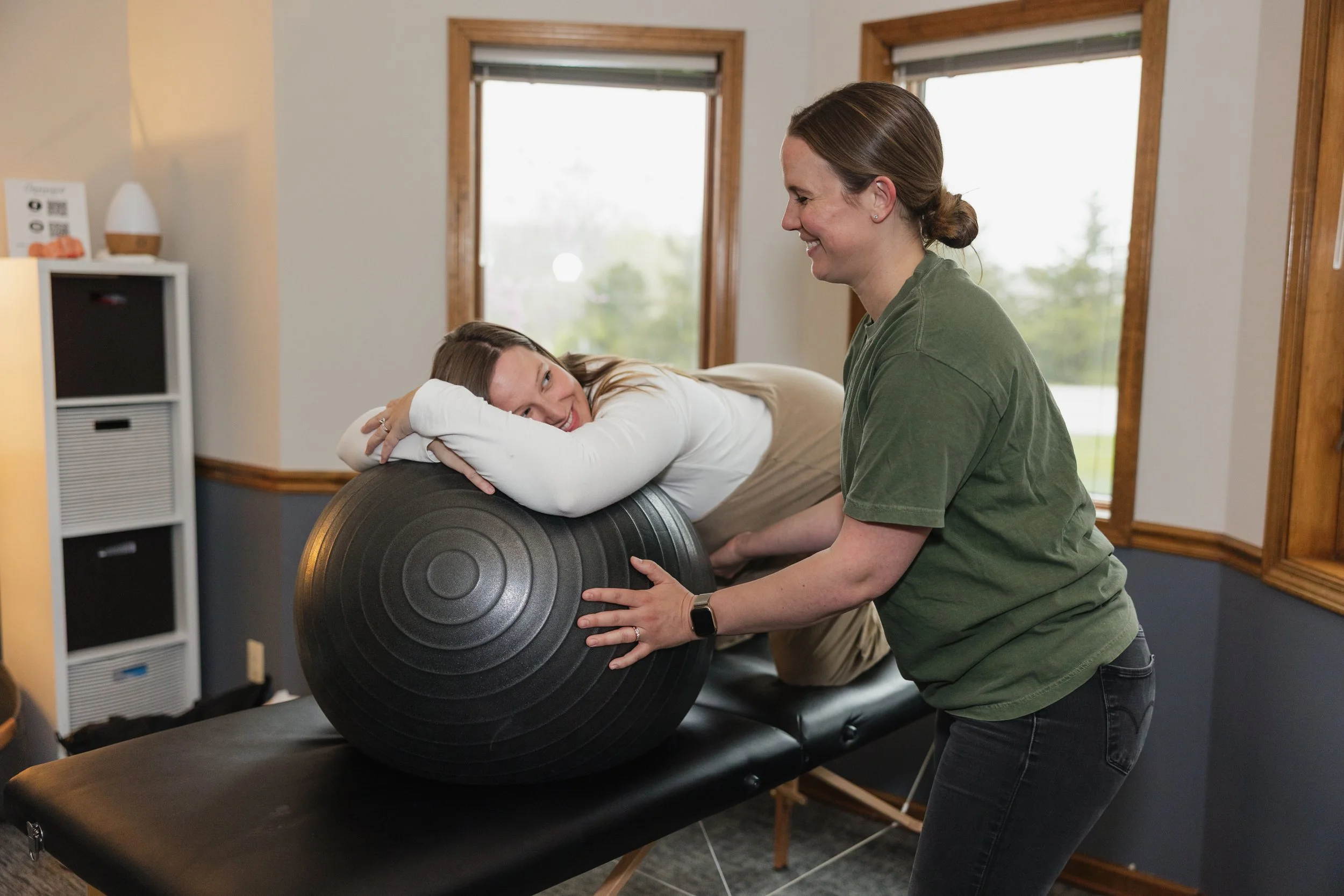 A woman lying on a treatment table rests her head on her arms while a therapist in a green shirt assists her with a large exercise ball during a physical therapy session in a room with windows.
