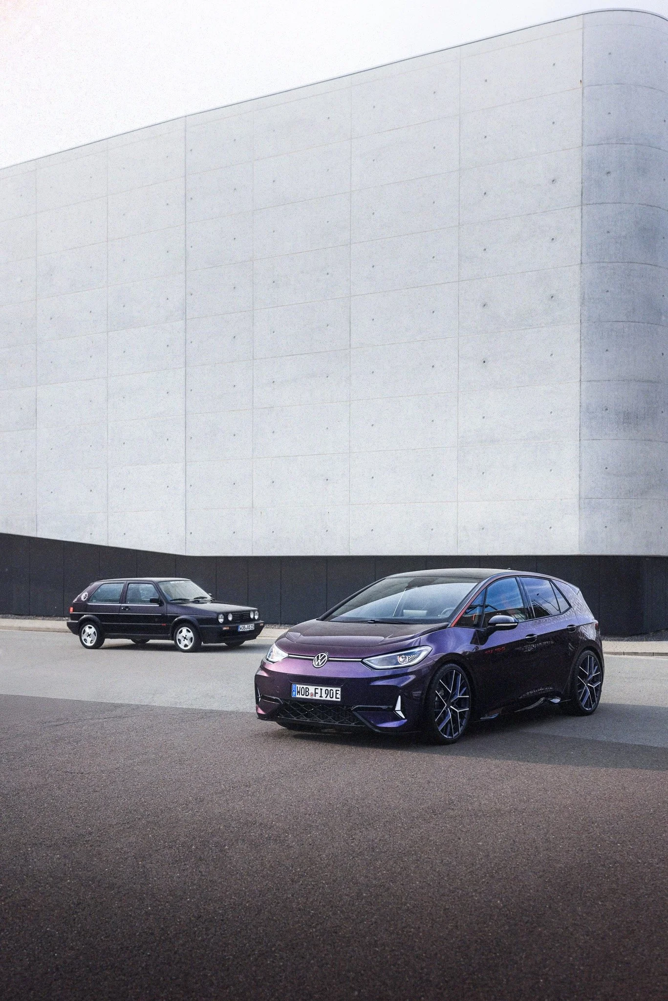 A purple Volkswagen car and a black older model hatchback car parked in front of a large modern concrete building