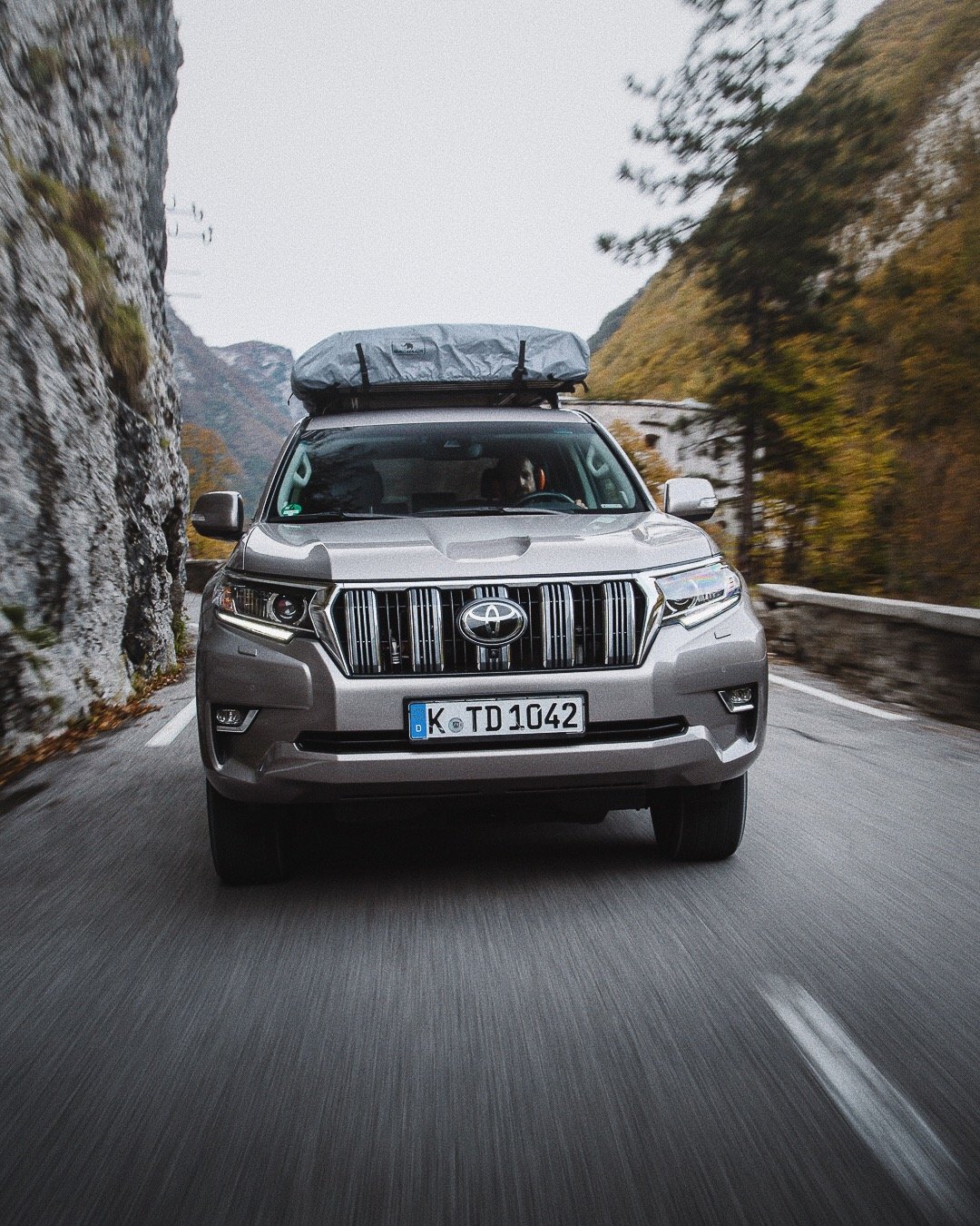 Silver Toyota SUV driving on a mountain road with autumn foliage on the sides, and a roof cargo bag on top.