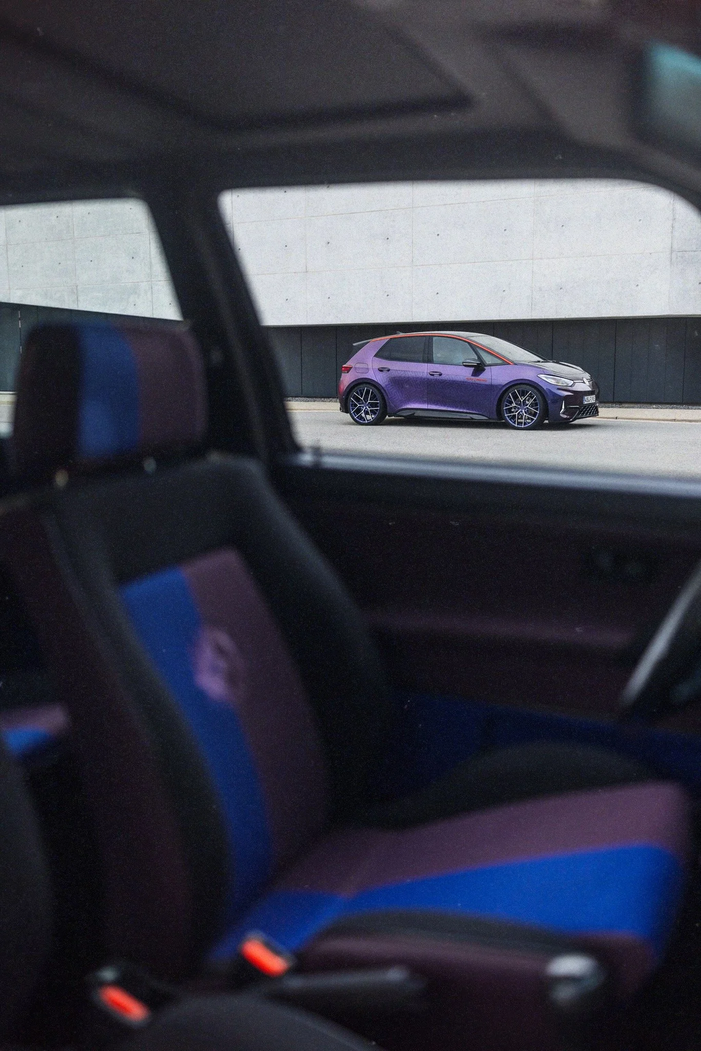 Photo taken from inside a car showing the side view of a purple hatchback parked on a street, with a modern building in the background.