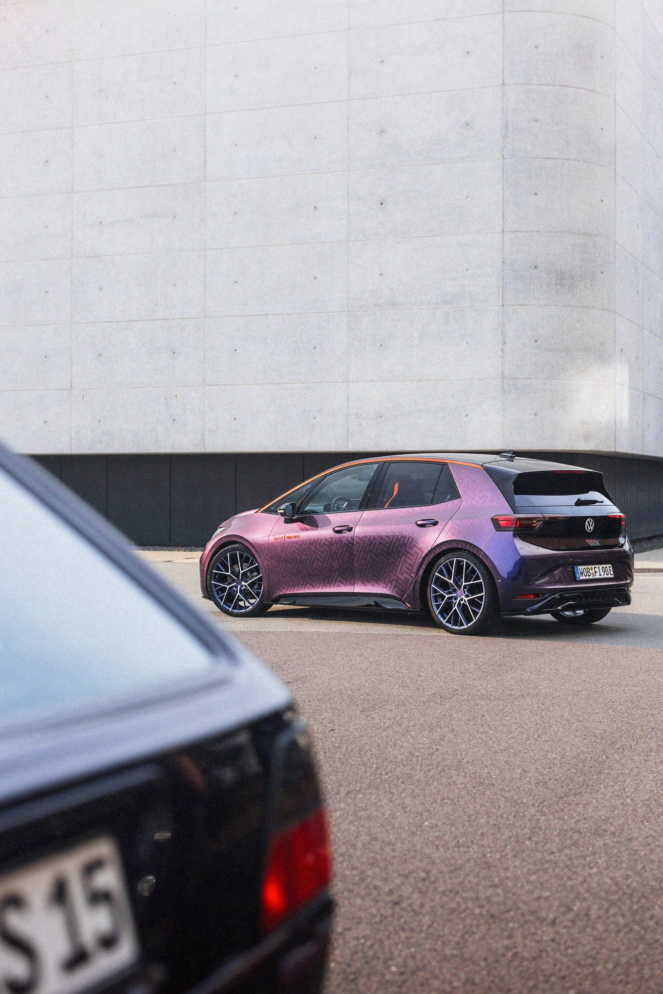 A purple and black Volkswagen vehicle parked on the street in front of a large concrete wall, with part of another car visible in the foreground.