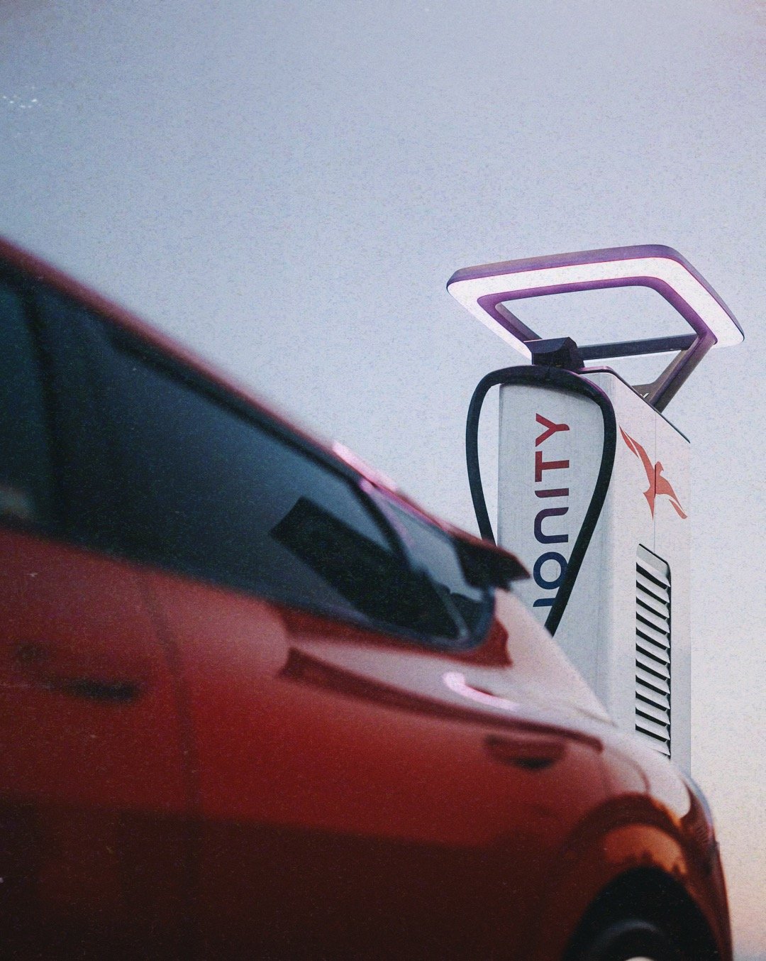 Close-up of a red car with a traffic camera or traffic enforcement device mounted on a pole in the background.