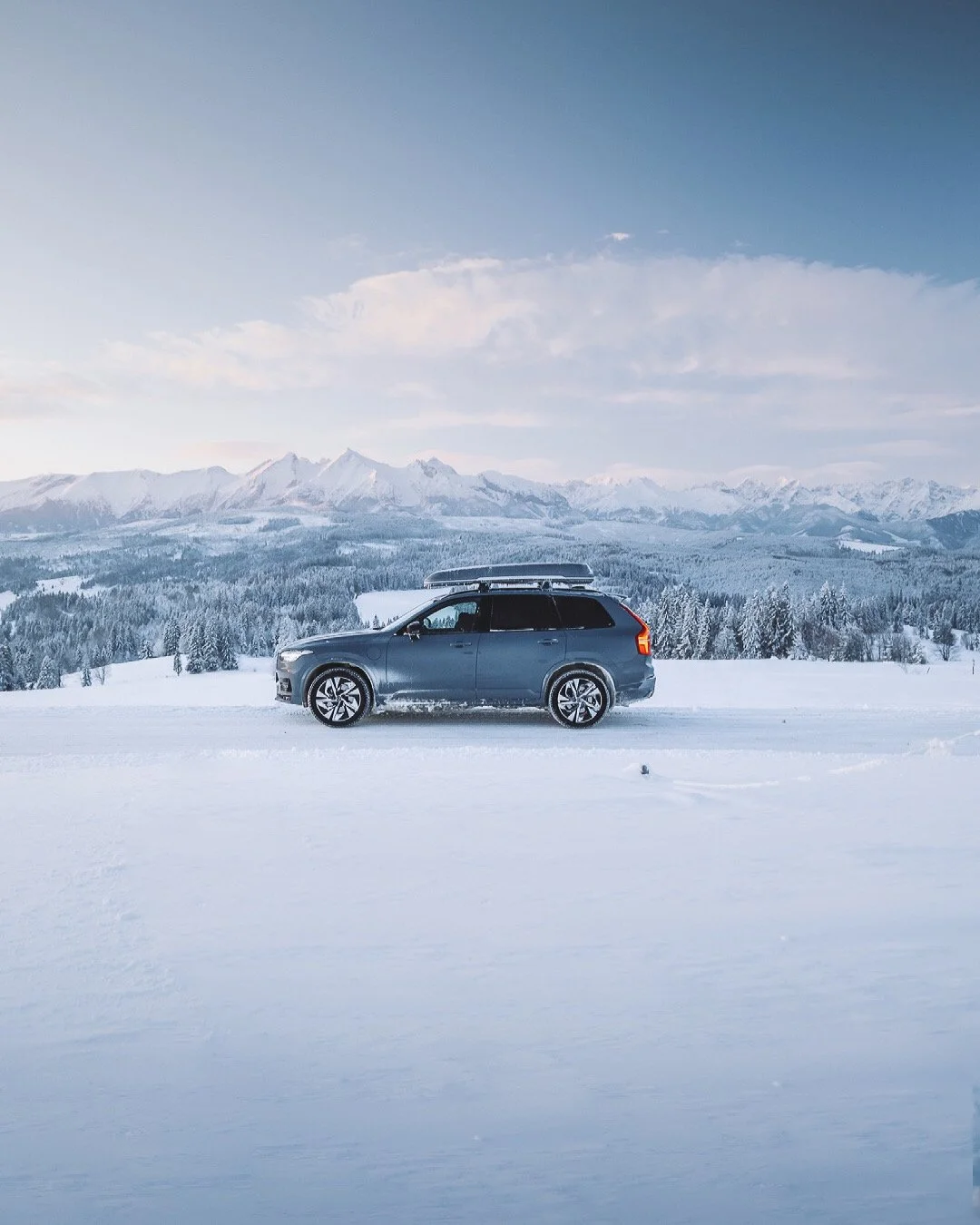 A dark gray station wagon with a roof cargo carrier parked on snow-covered ground in a winter landscape with snow-covered trees and mountain range in the background.