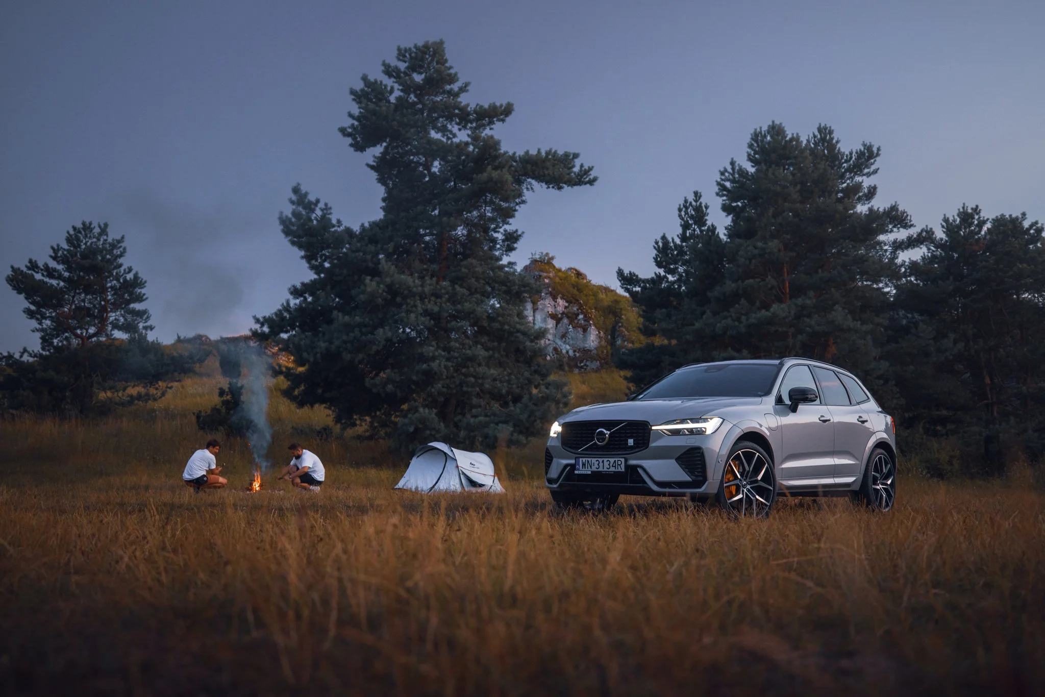 Two people camping on a grassy field at dusk, sitting around a small campfire with smoke rising, near a gray Volvo SUV with a European license plate, and a small tent set up among tall pine trees in the background.