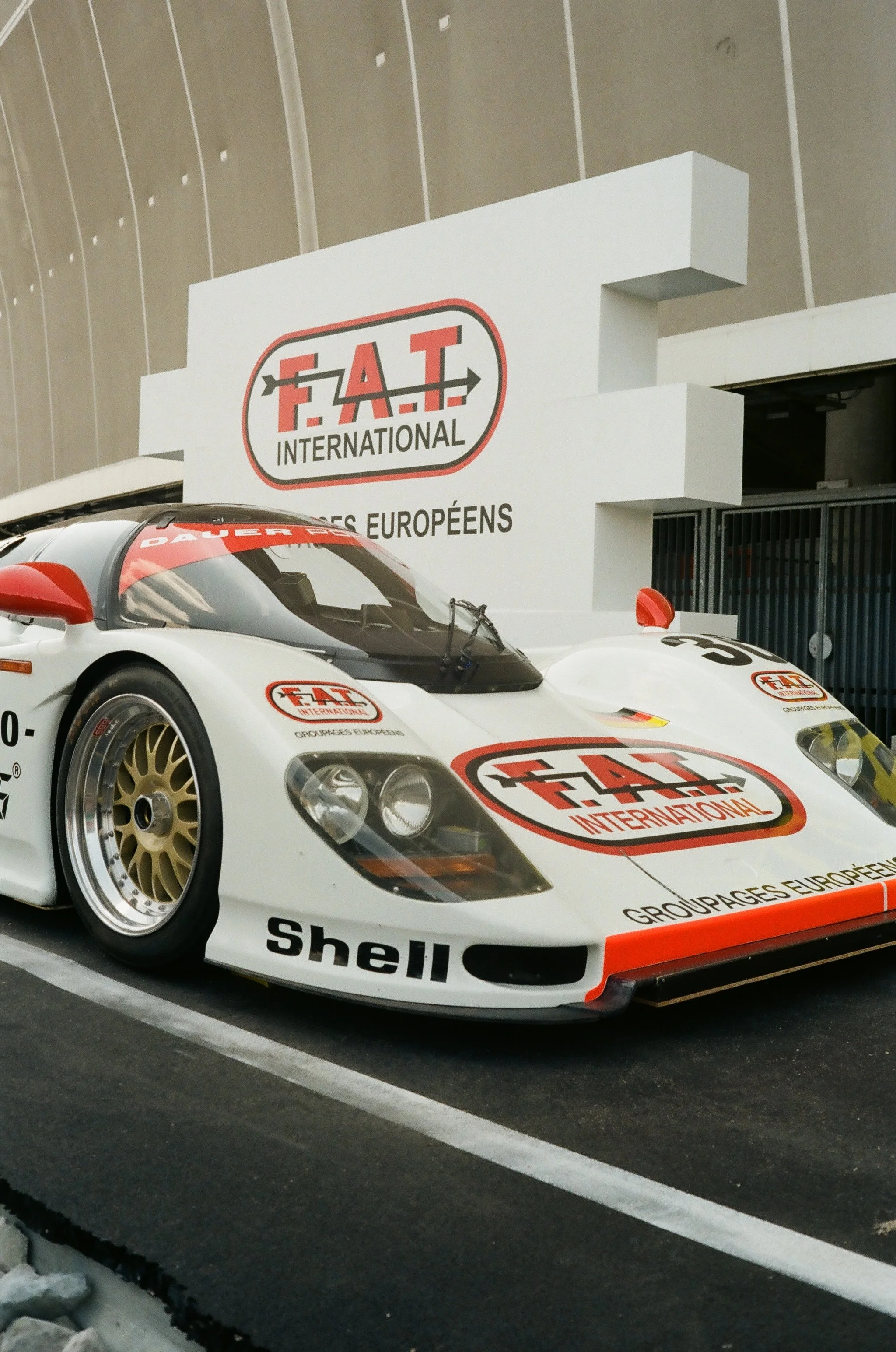 White race car with Ferrari logo, red accents, and sponsor decals, parked in front of a sign that reads 'F.A.T International' at a parking lot.
