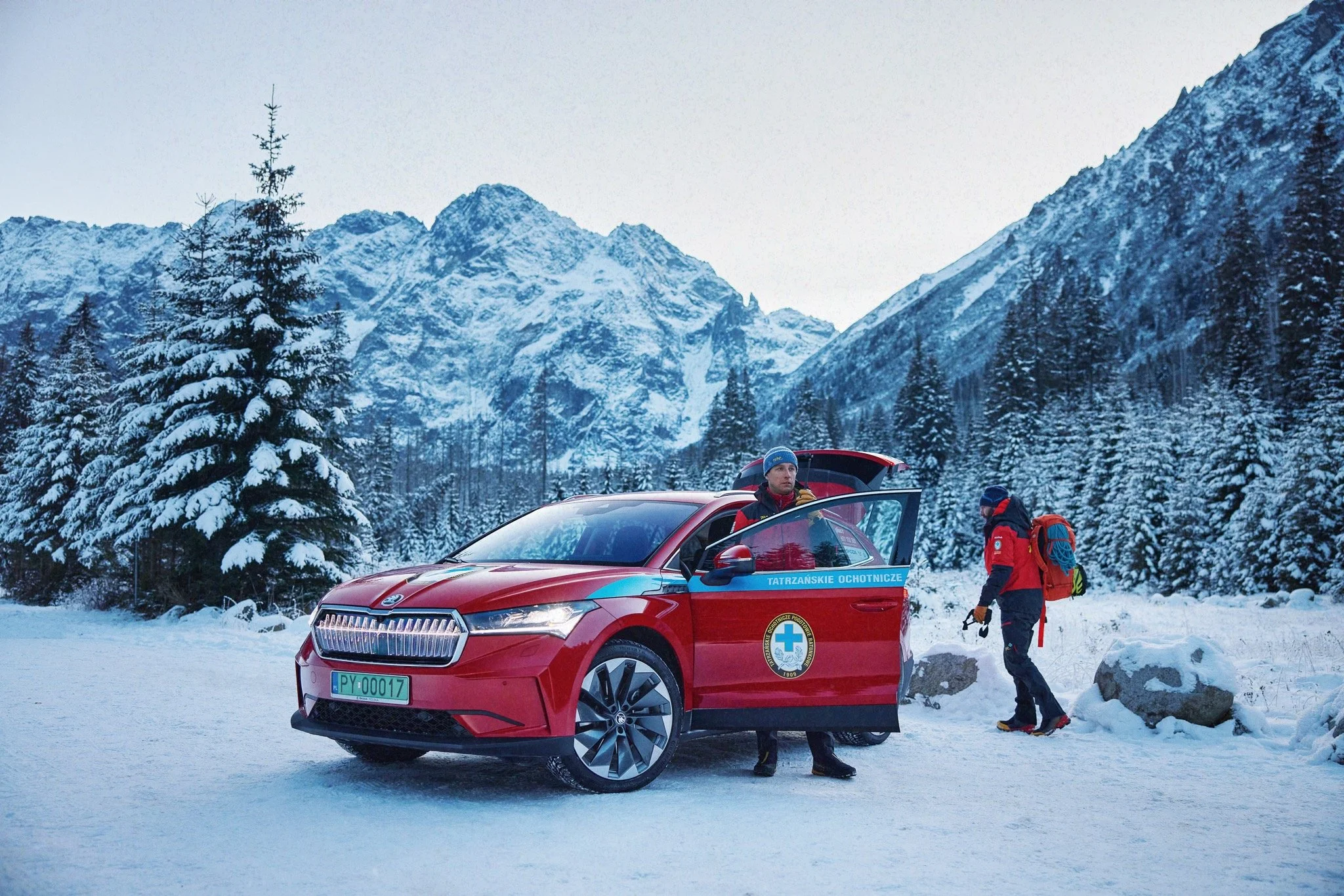 Two rescue workers with a red vehicle in a snowy mountain landscape, one standing by the car and the other walking nearby, surrounded by snow-covered trees and mountains.