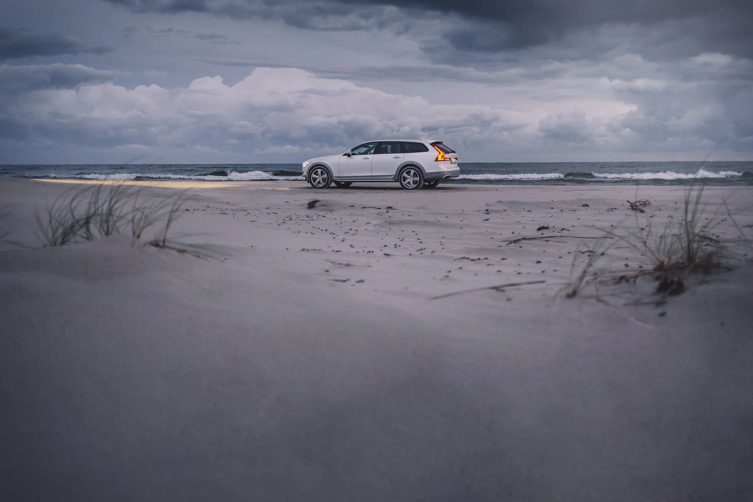 A white station wagon parked on a sandy beach near the ocean with cloudy skies overhead.