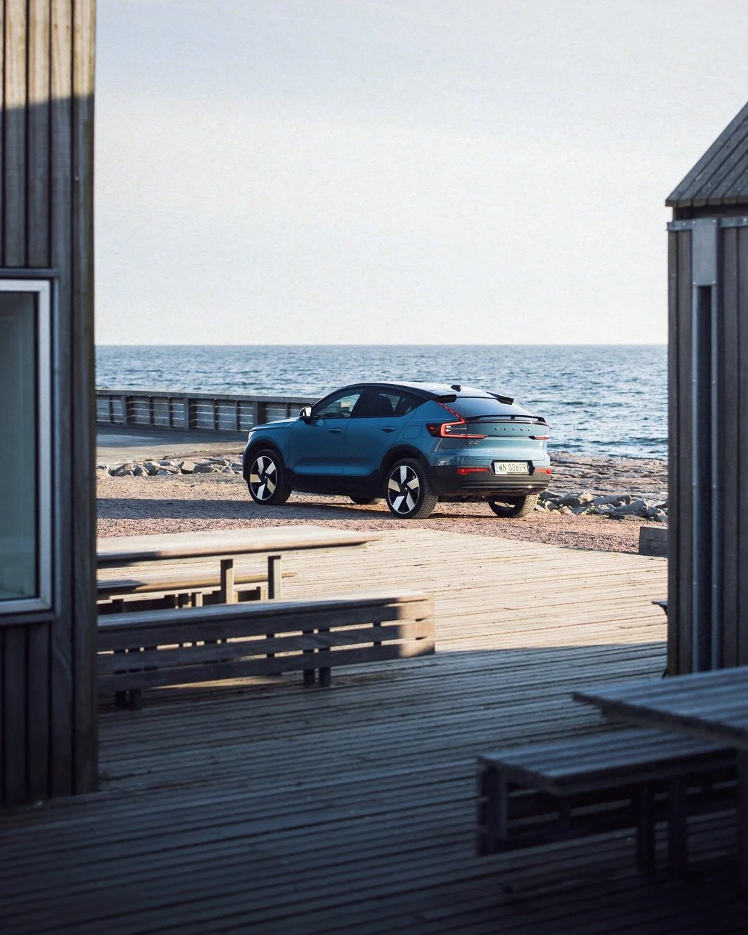Blue car parked on a beach in view from a seaside cabin with wooden decks and benches, overlooking the ocean.