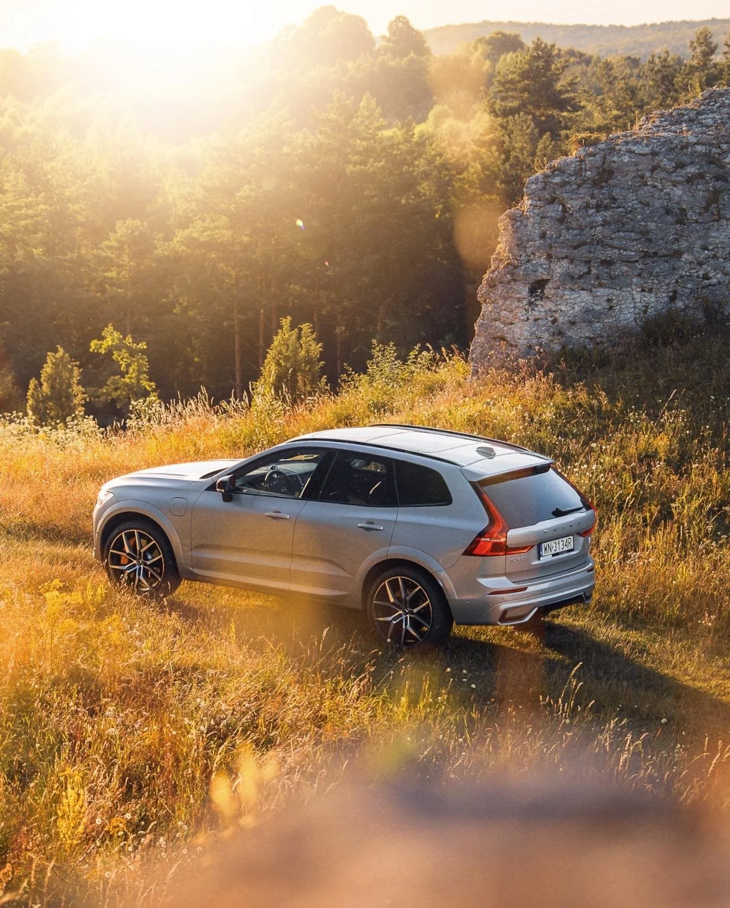 A silver SUV parked in a grassy field with a rocky cliff and a forest in the background during sunset.