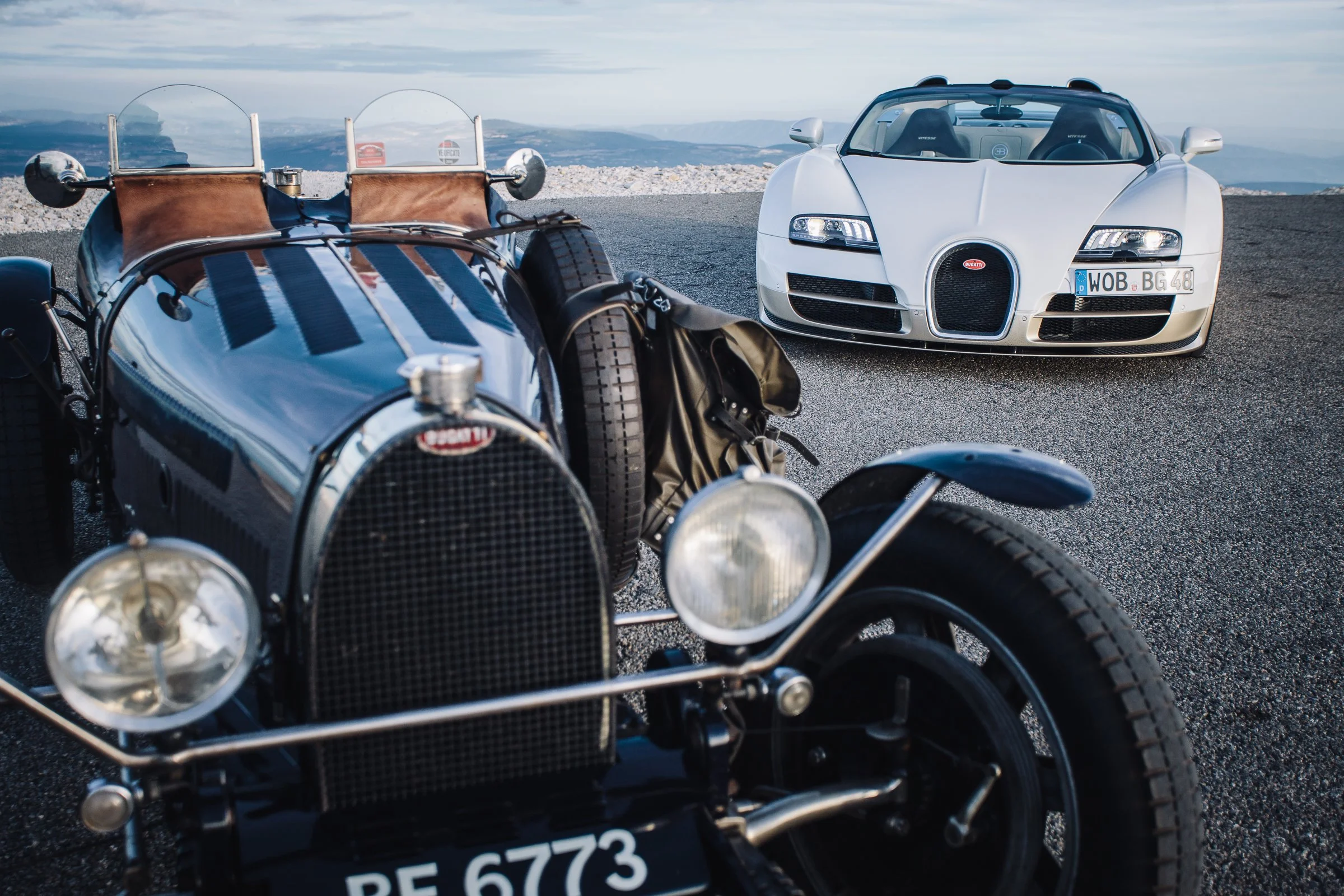 A vintage black racing car with a leather seat and chrome accents, alongside a modern white Bugatti supercar, both on a mountain road with a scenic cloudy sky and distant mountains in the background.