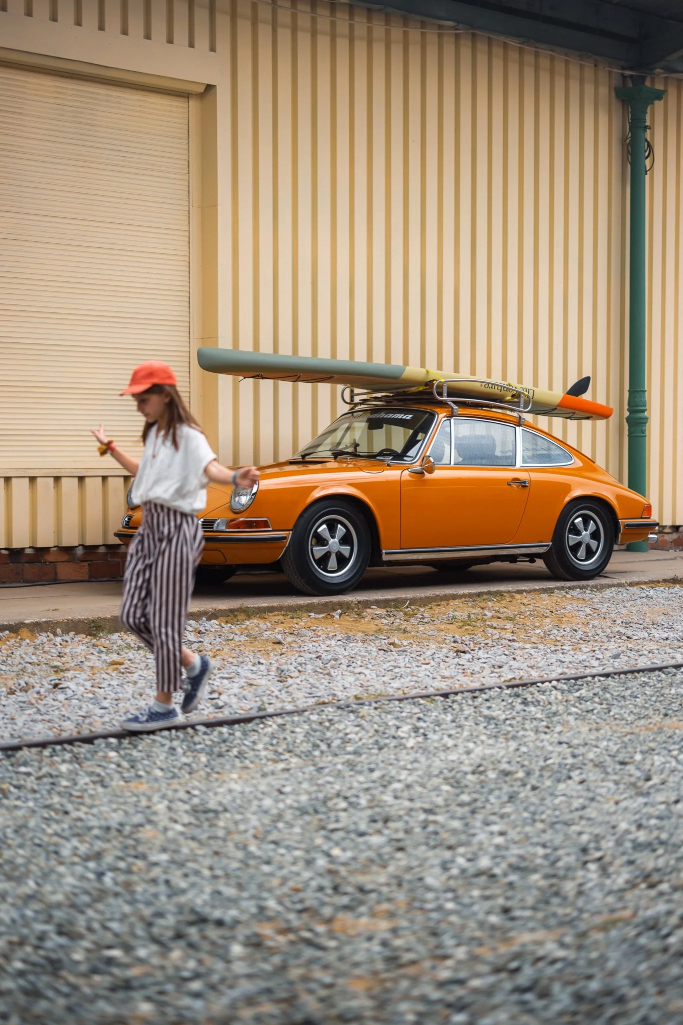 A vintage orange Porsche 911 with a surfboard on top, parked beside a yellow building with vertical stripes. A girl in striped pants, a white shirt, and a red cap walks in front of the car, gesturing with one hand.