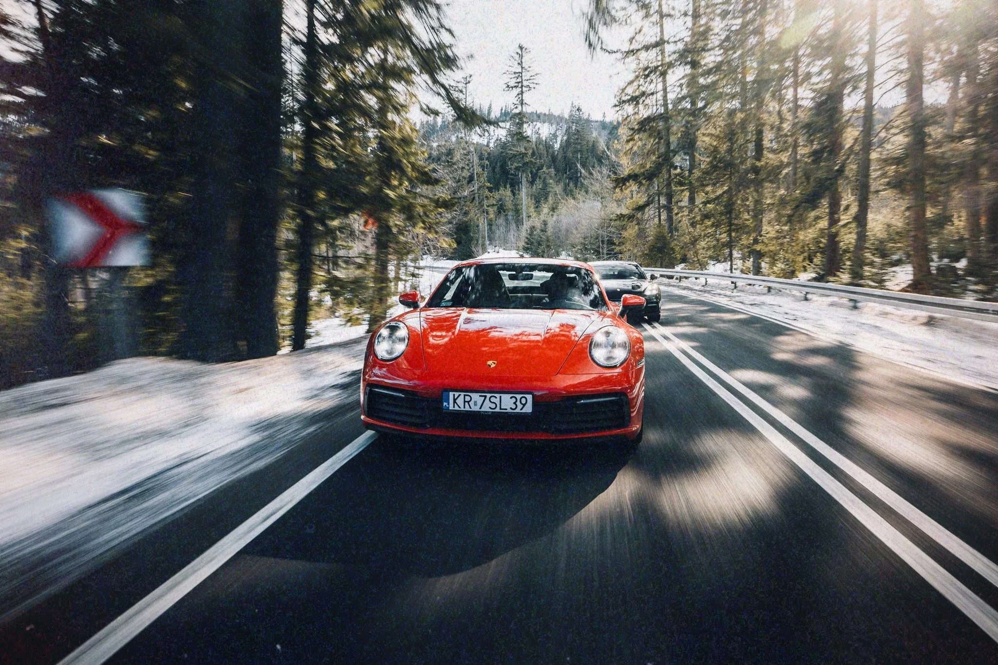 Red Porsche sports car driving on a winding mountain road through a snowy forest during daylight.