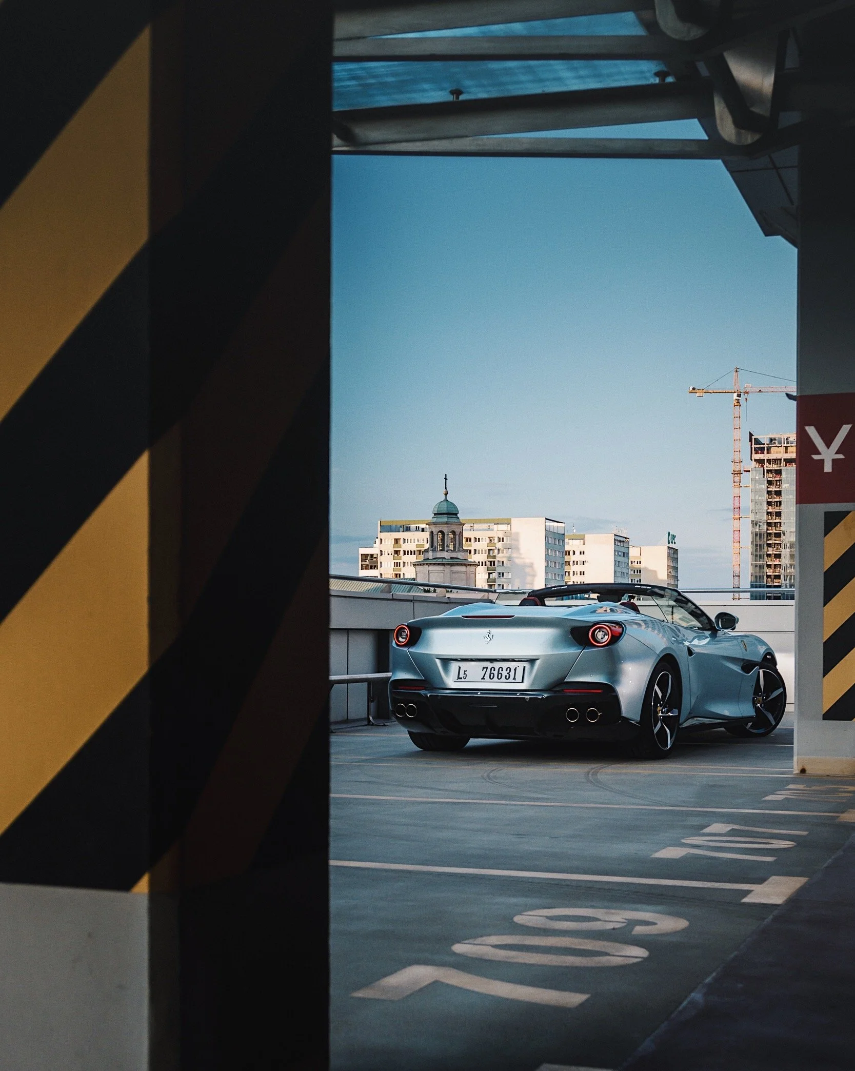 Silver sports car parked on the top level of a parking garage with city buildings and a crane in the background, framed by black and yellow striped barriers.