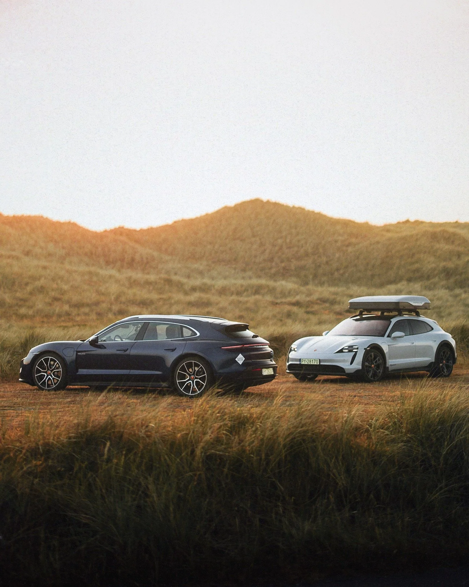 Two modern cars parked on a grassy field at sunset, with rolling hills in the background.