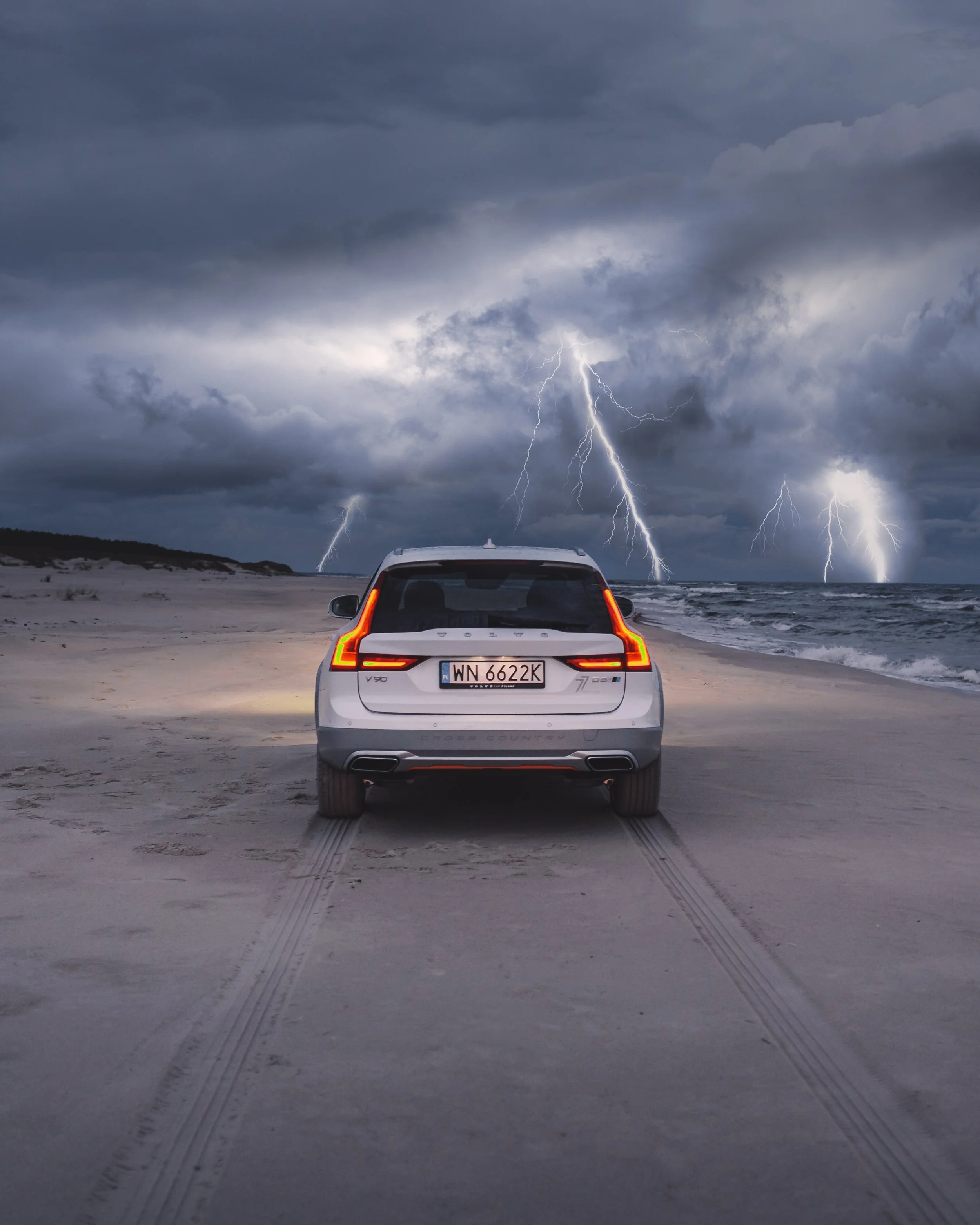 White Volvo SUV parked on a beach during a thunderstorm with lightning strikes in the cloudy sky.