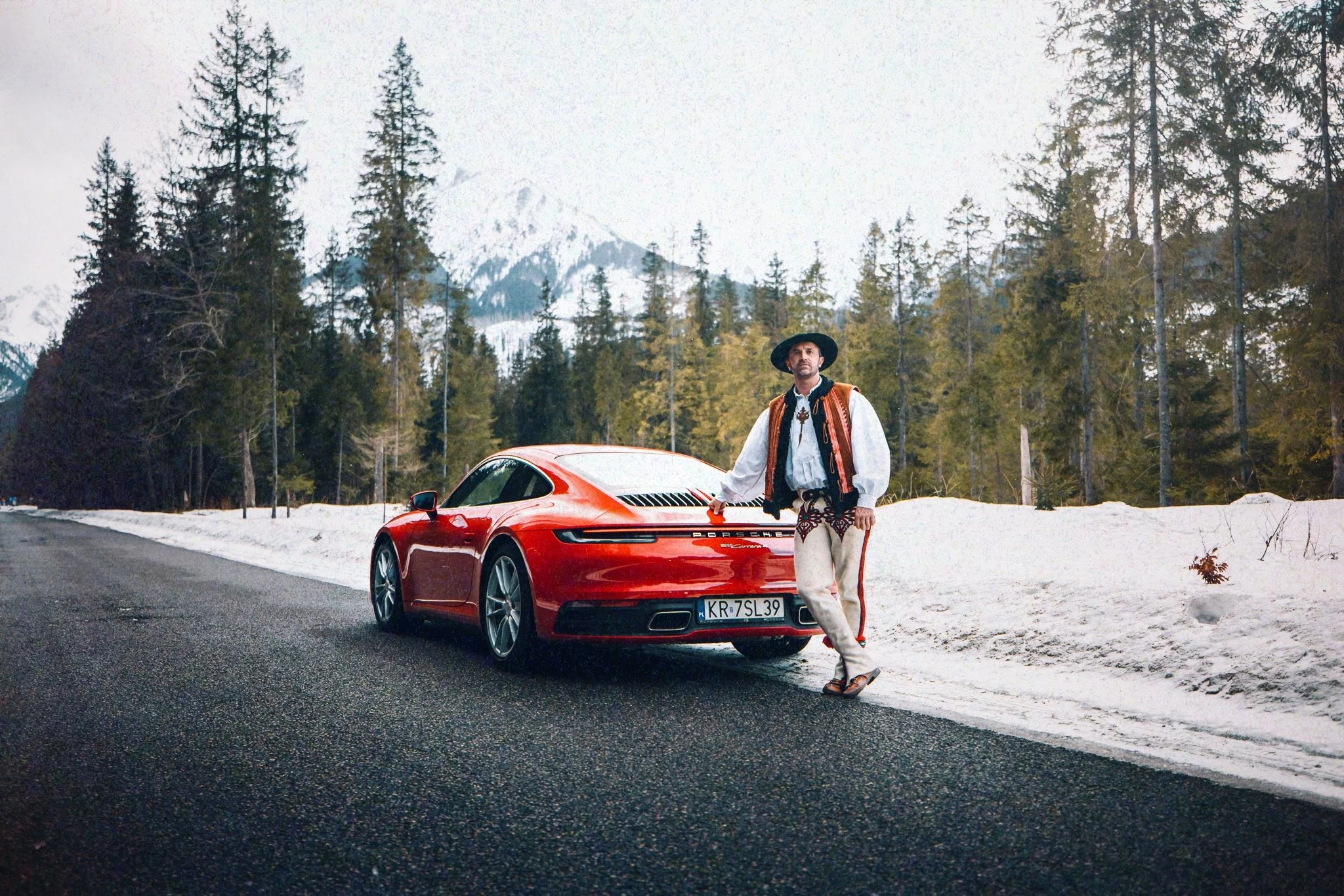 A man dressed in traditional clothing standing next to a red Porsche car on a snowy mountain road, with pine trees and snow-covered mountains in the background.