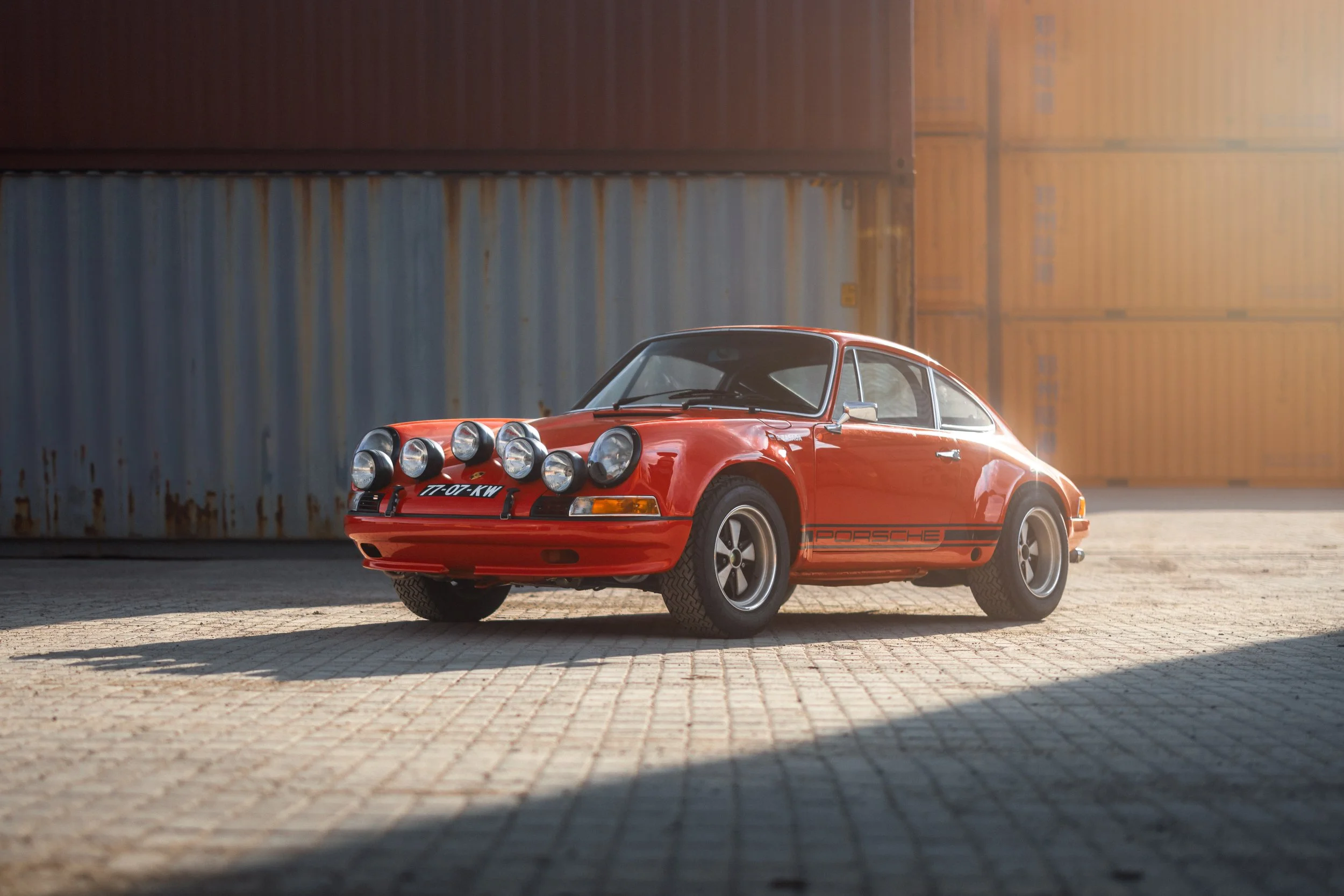 A red vintage Porsche 911 with rally lights on the front, parked on a paved lot with shipping containers in the background and sunlight creating a warm glow.