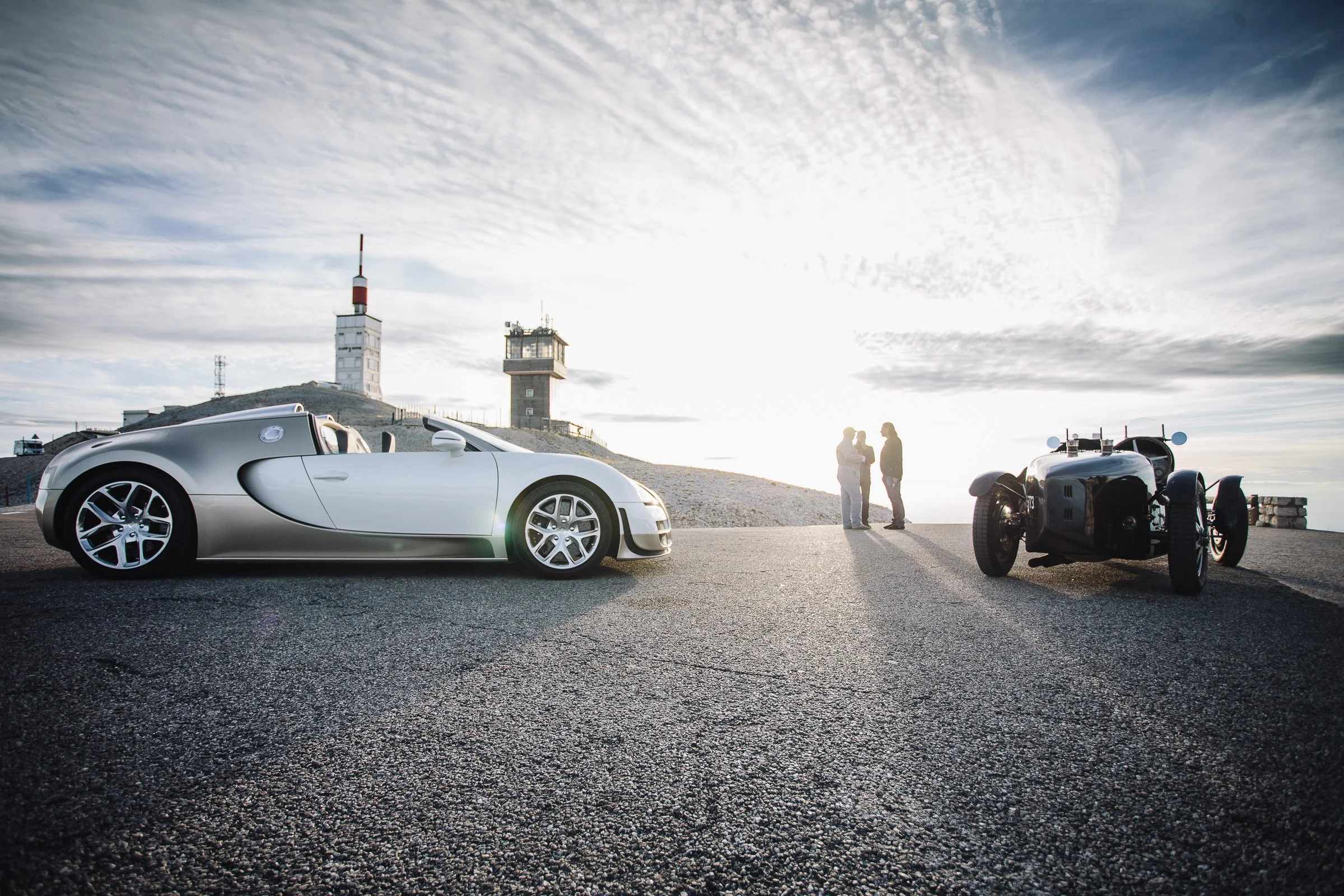 A silver sports car and a black vintage race car parked on a hilltop with two people talking in the background during sunset.
