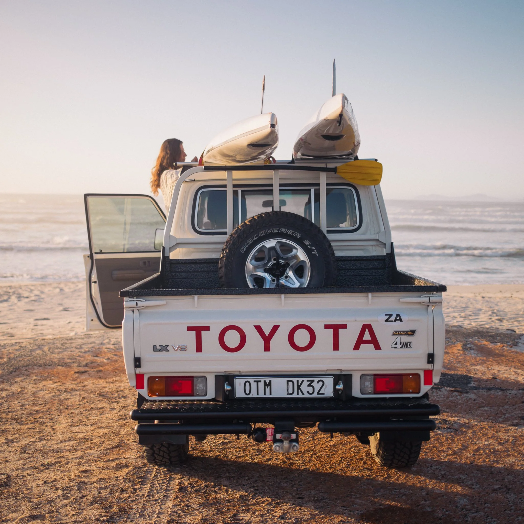 White Toyota pickup truck parked on the beach with two kayaks and a woman standing outside, looking at the ocean during sunset.