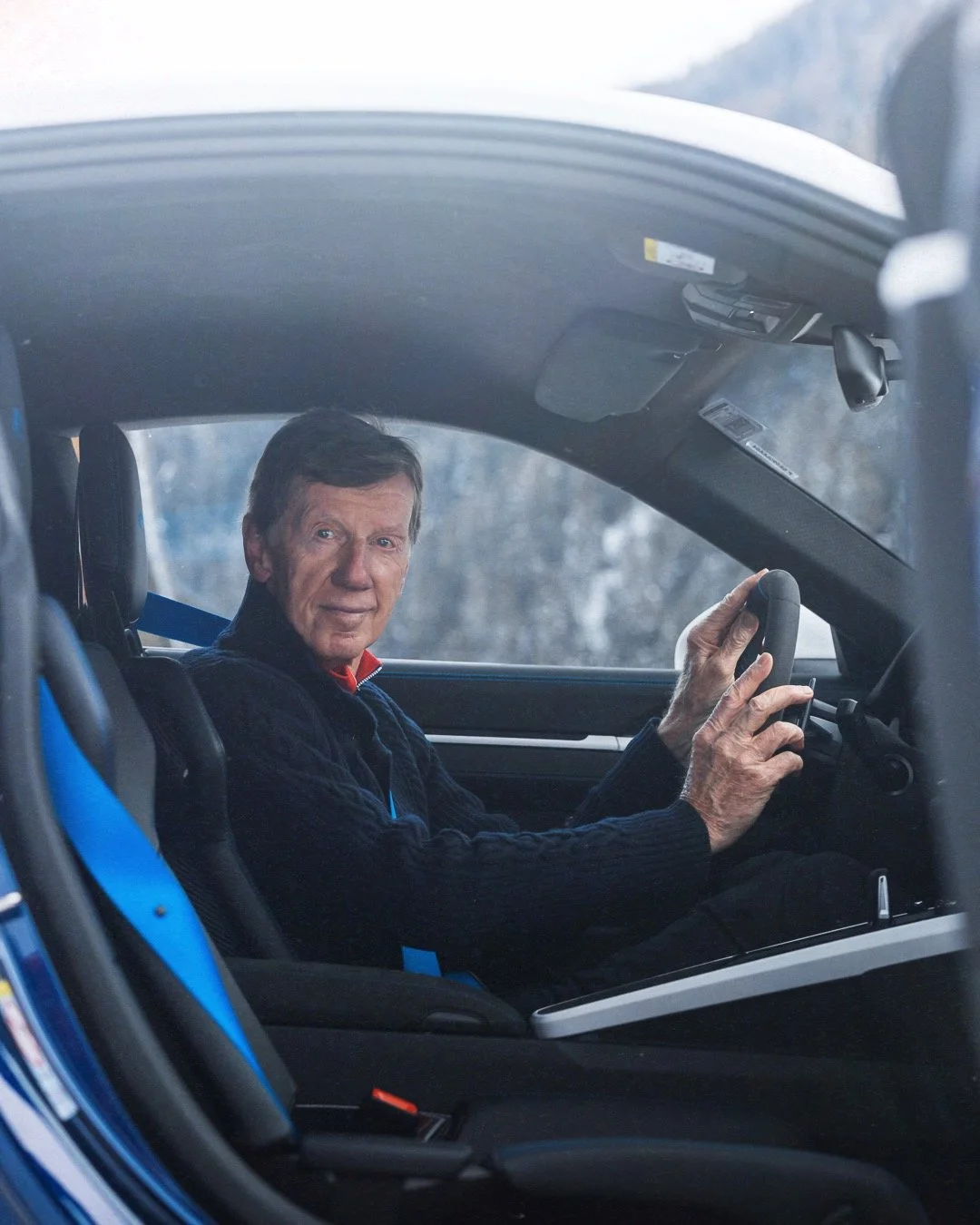 An elderly man sitting in a car, holding the steering wheel, looking at the camera.