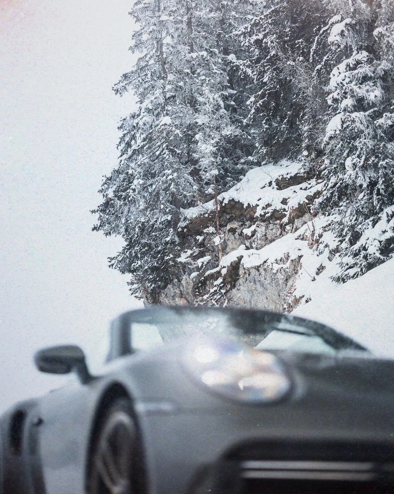 View through a car windshield of a snow-covered mountain road with tall pine trees and a rocky cliffside.