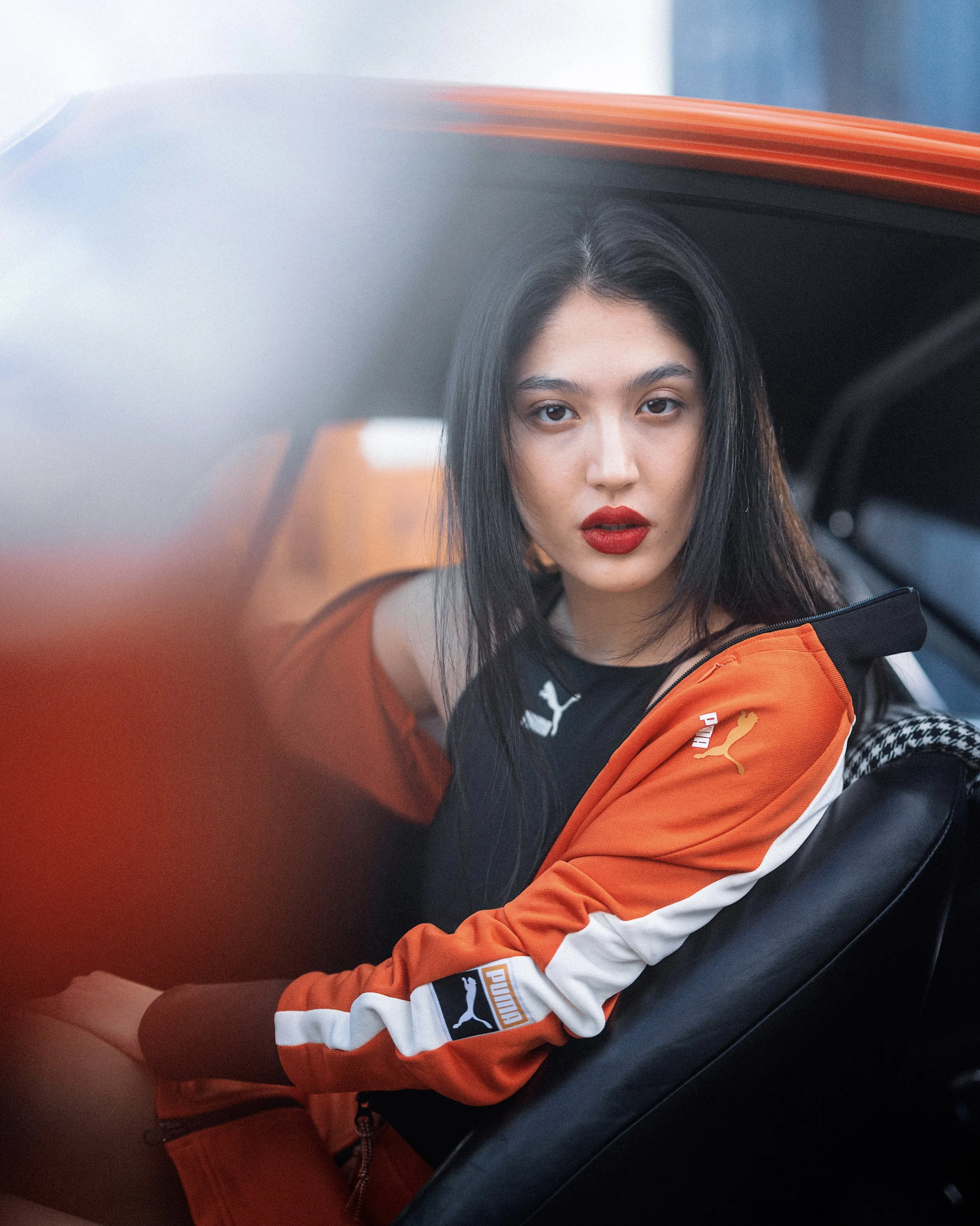 Young woman with long black hair, red lipstick, wearing an orange and black Puma jacket, sitting in a car.