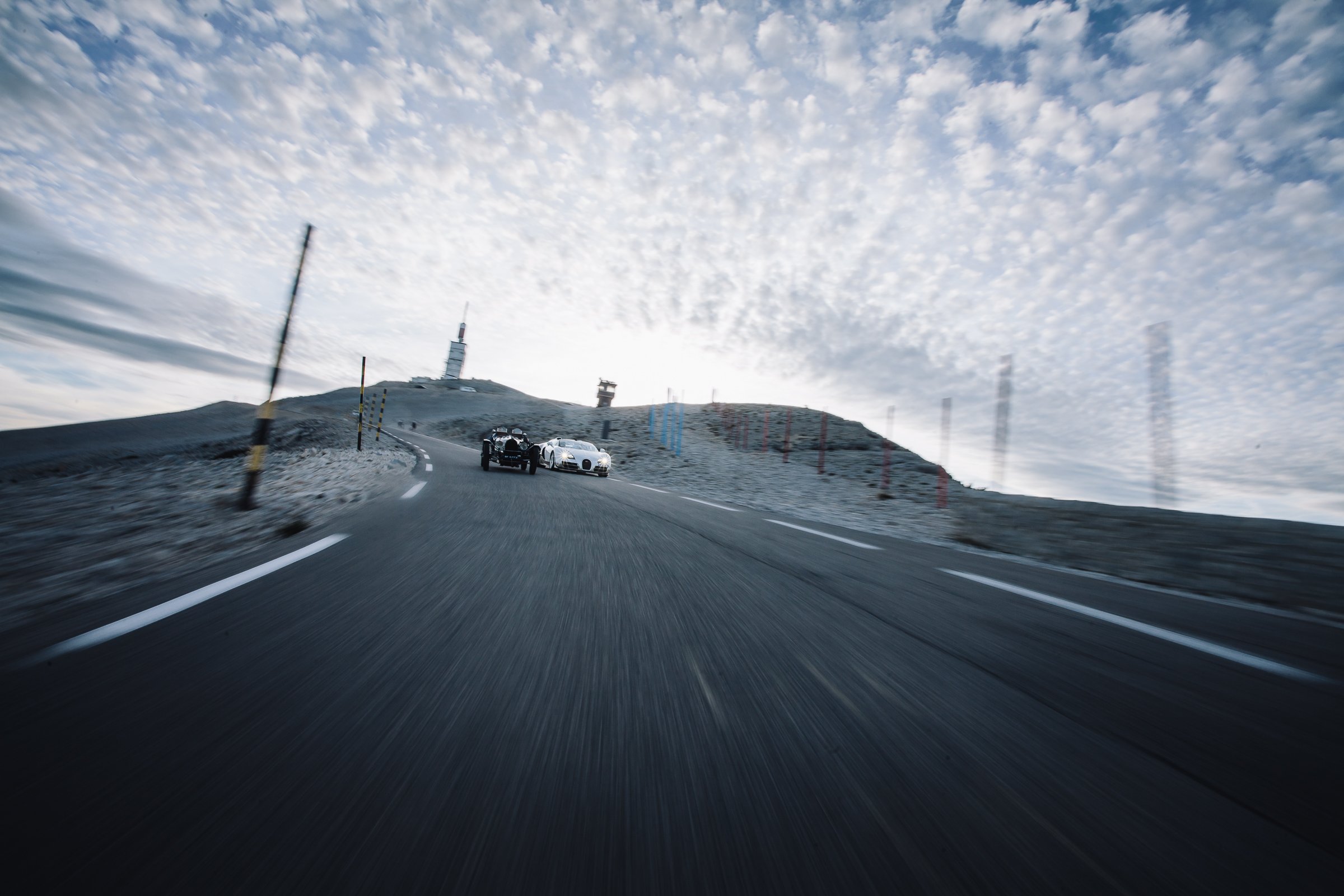 Two cars driving on a mountain road under a cloudy sky with an antenna tower on the peak in the background.