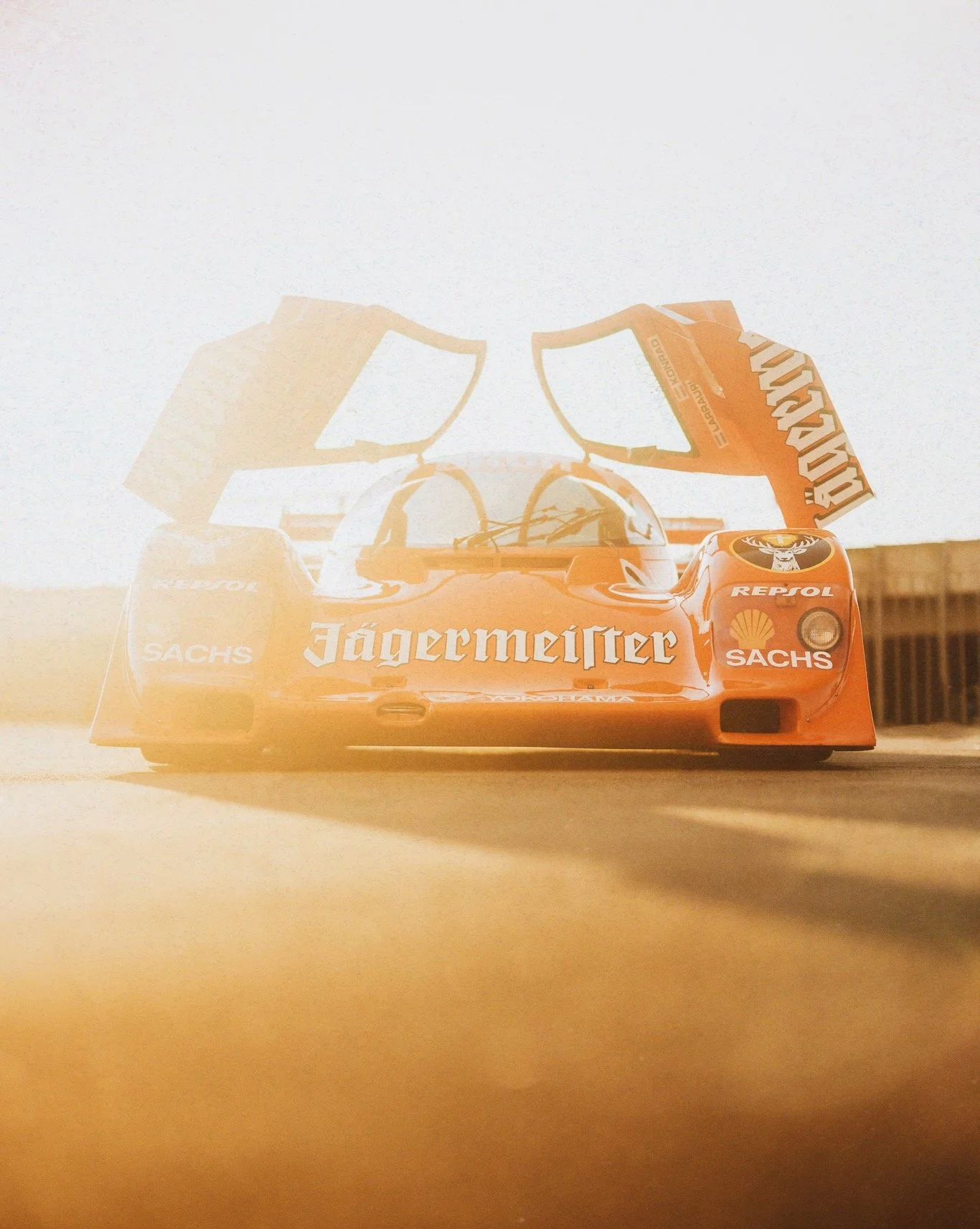 A vintage race car on a racetrack with its rear wing raised, displaying sponsor logos including Jägermeister, Repsol, Shell, and Sachs, illuminated by bright sunlight.