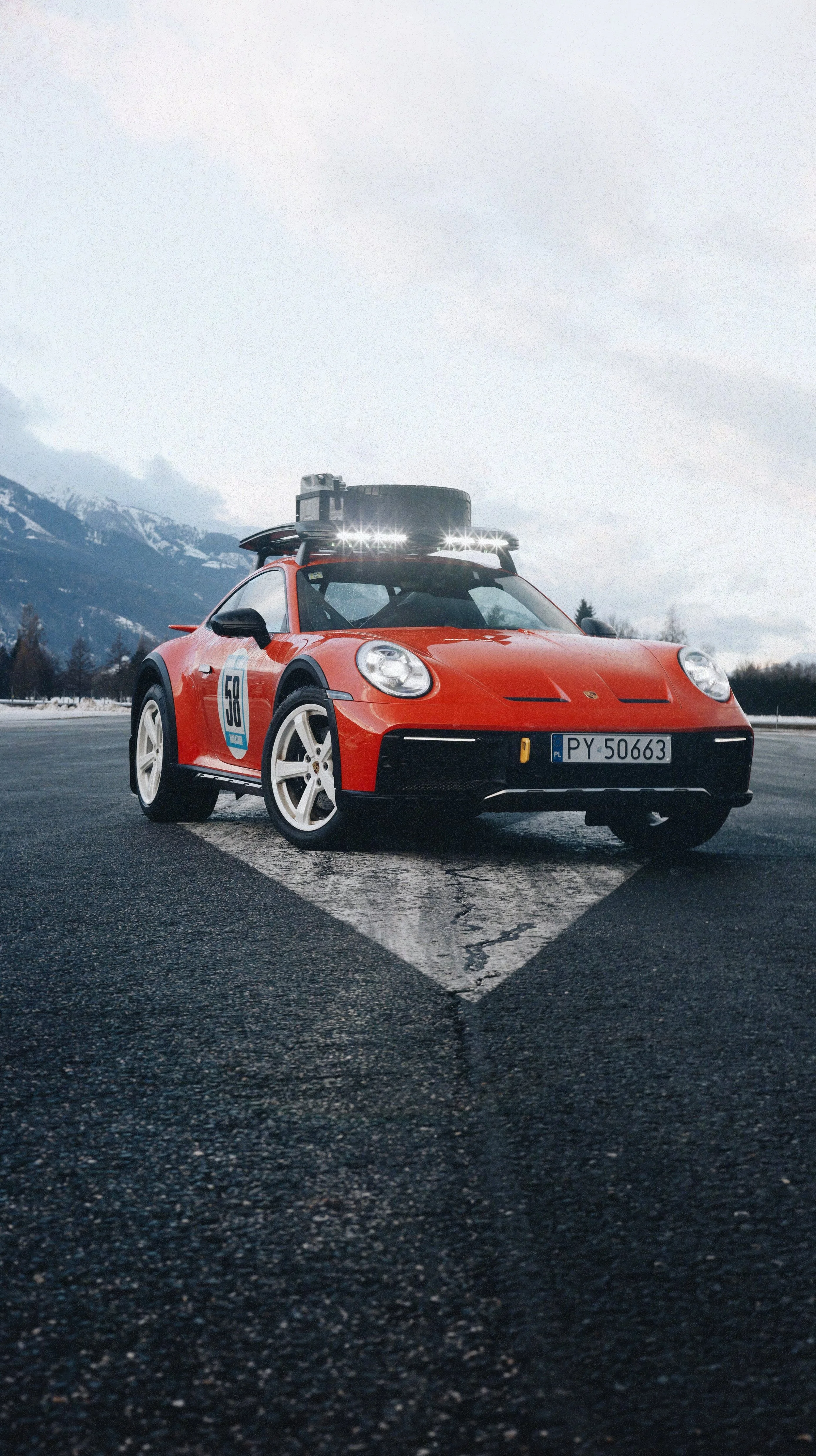 A red Porsche sports car with a racing number on the door, parked on an empty road with snowy mountains in the background and an overcast sky.