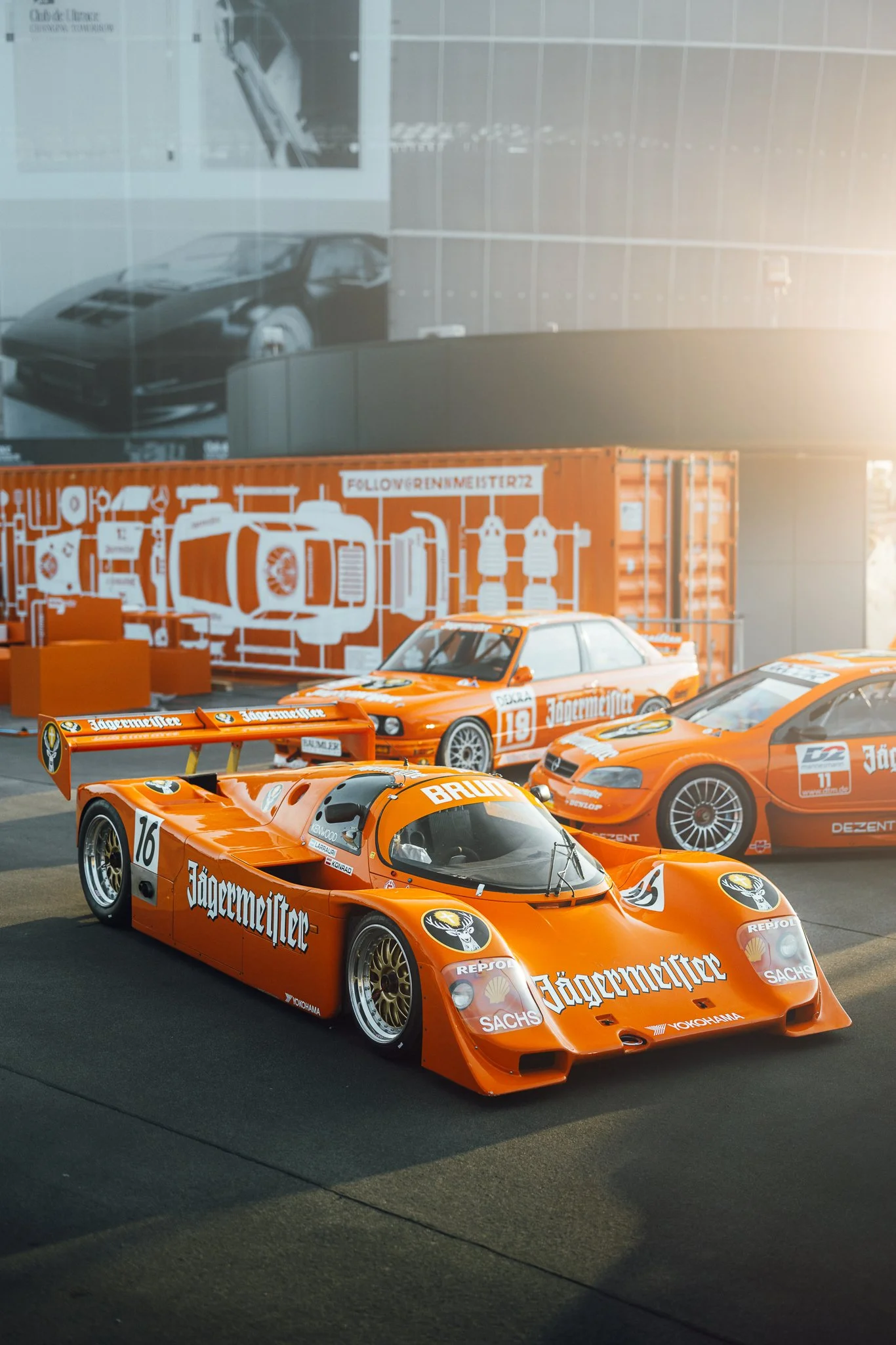 A vintage orange race car with Jägermeister sponsorship, number 16, parked on display with other race cars and memorabilia in the background.