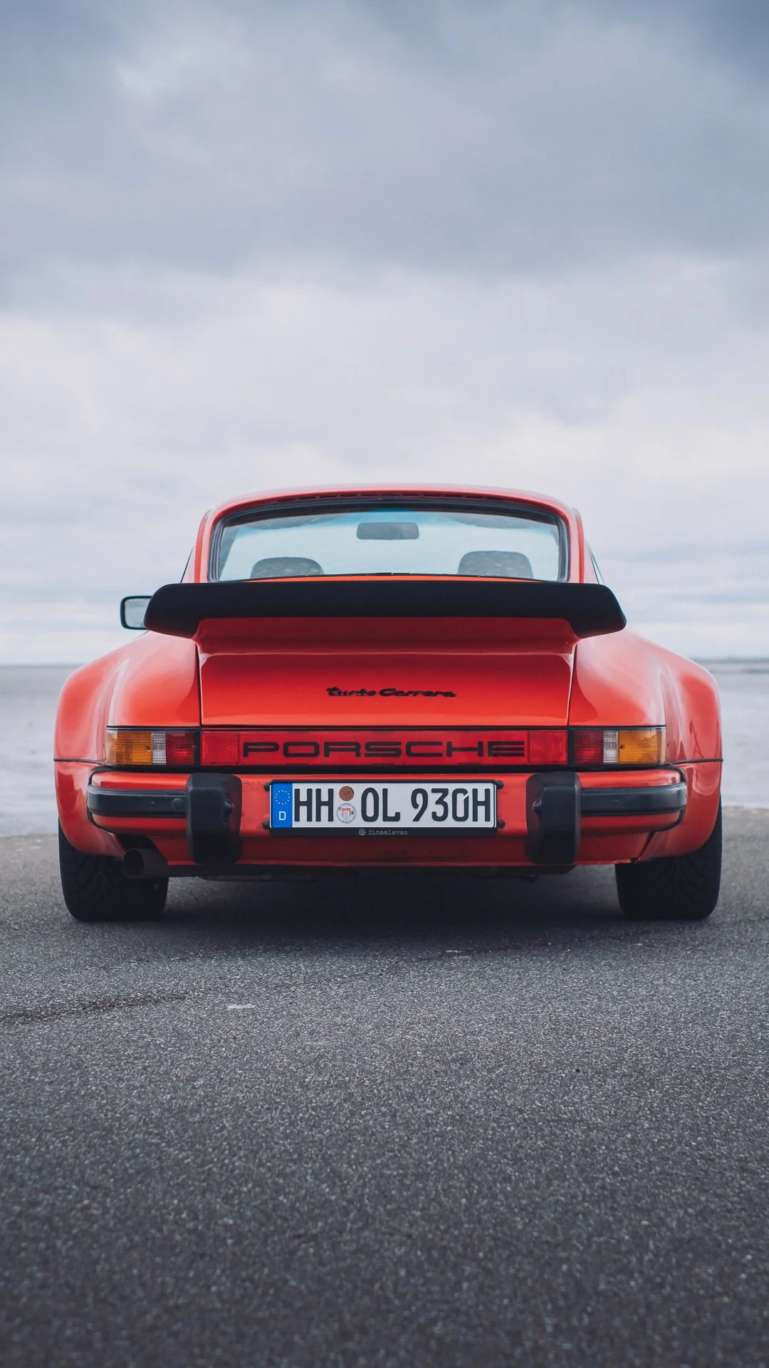 Rear view of a classic red Porsche Turbo Carrera sports car parked on a paved surface near water, with a cloudy sky in the background.