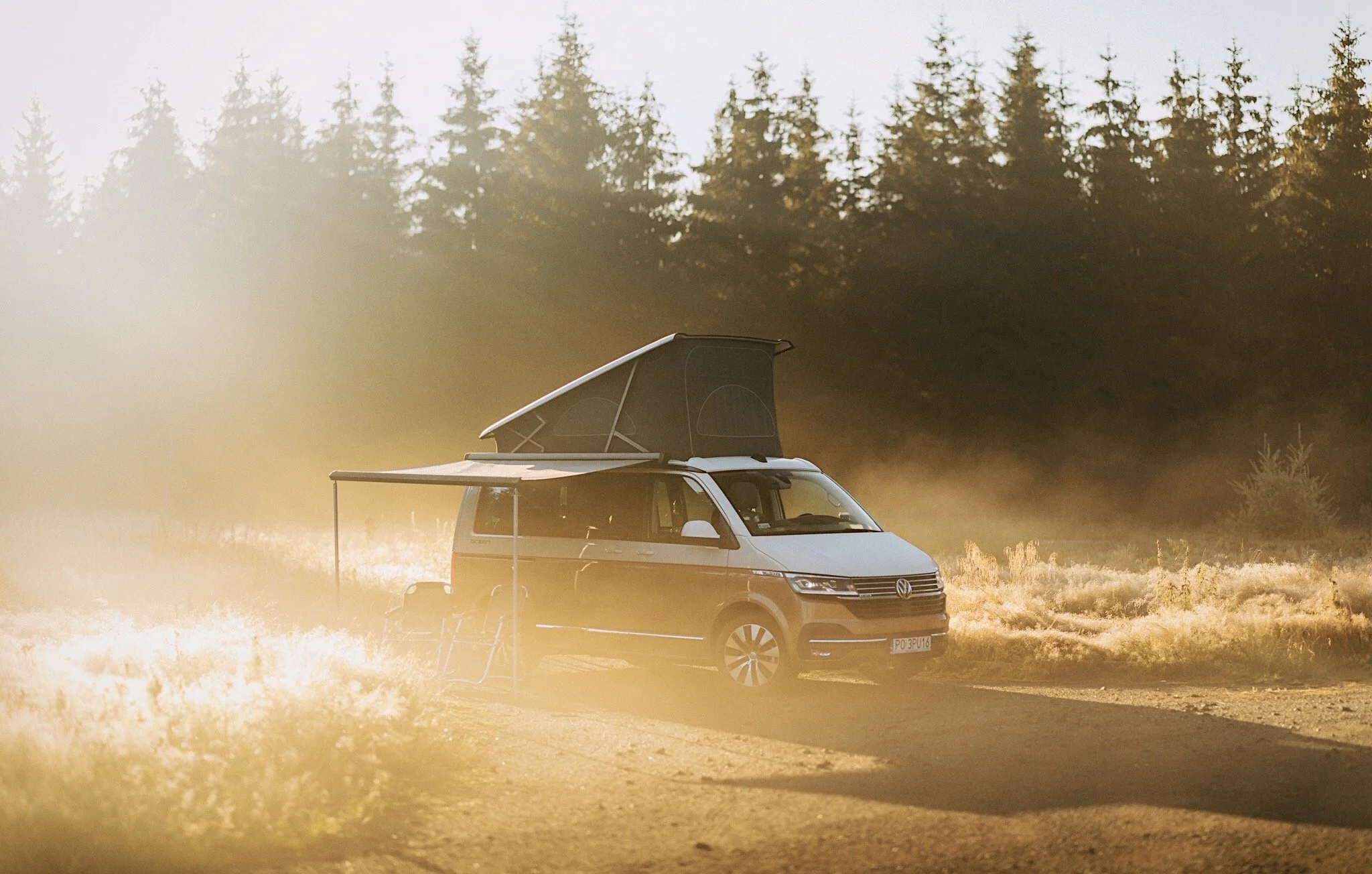 A camper van on a dirt road in a forested area during sunset, with a pop-up roof tent extended and a foldable chair beside it.