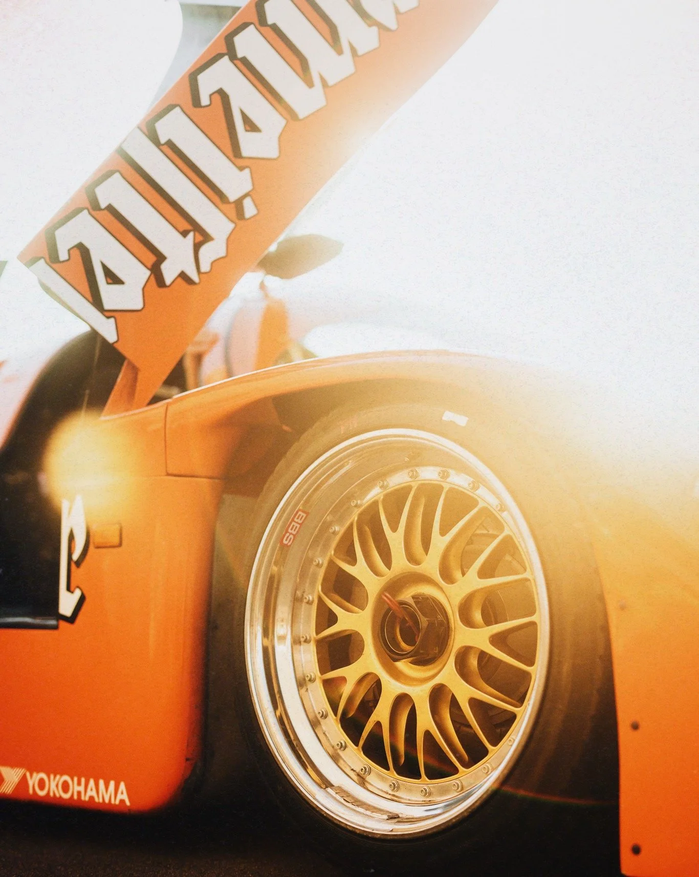 Close-up of an orange race car with Yokohama branding and a gold BBS wheel showing the center cap.