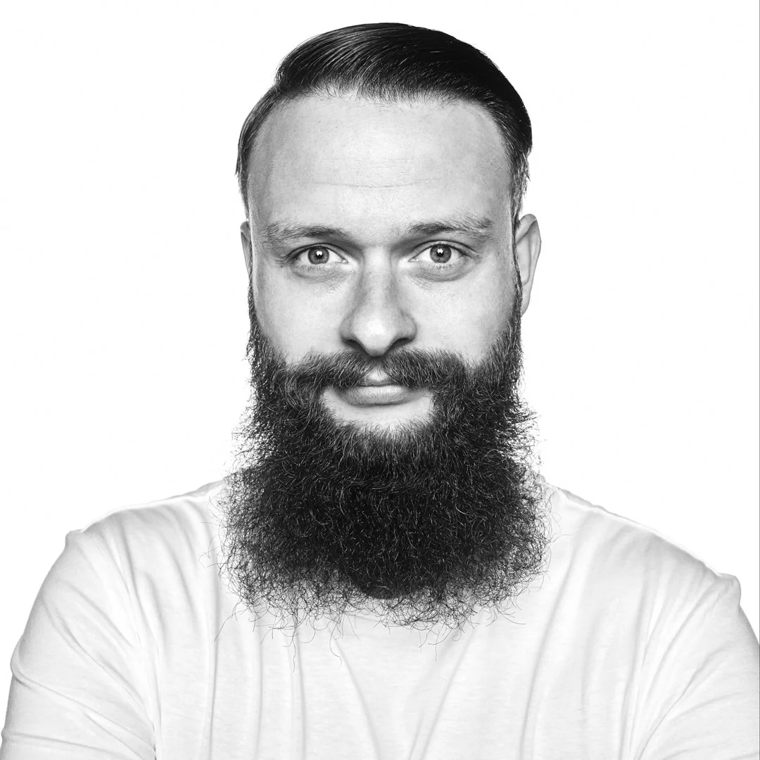 Black and white portrait of a man with a well-groomed beard, mustache, short hair, and wearing a white shirt, against a plain background.