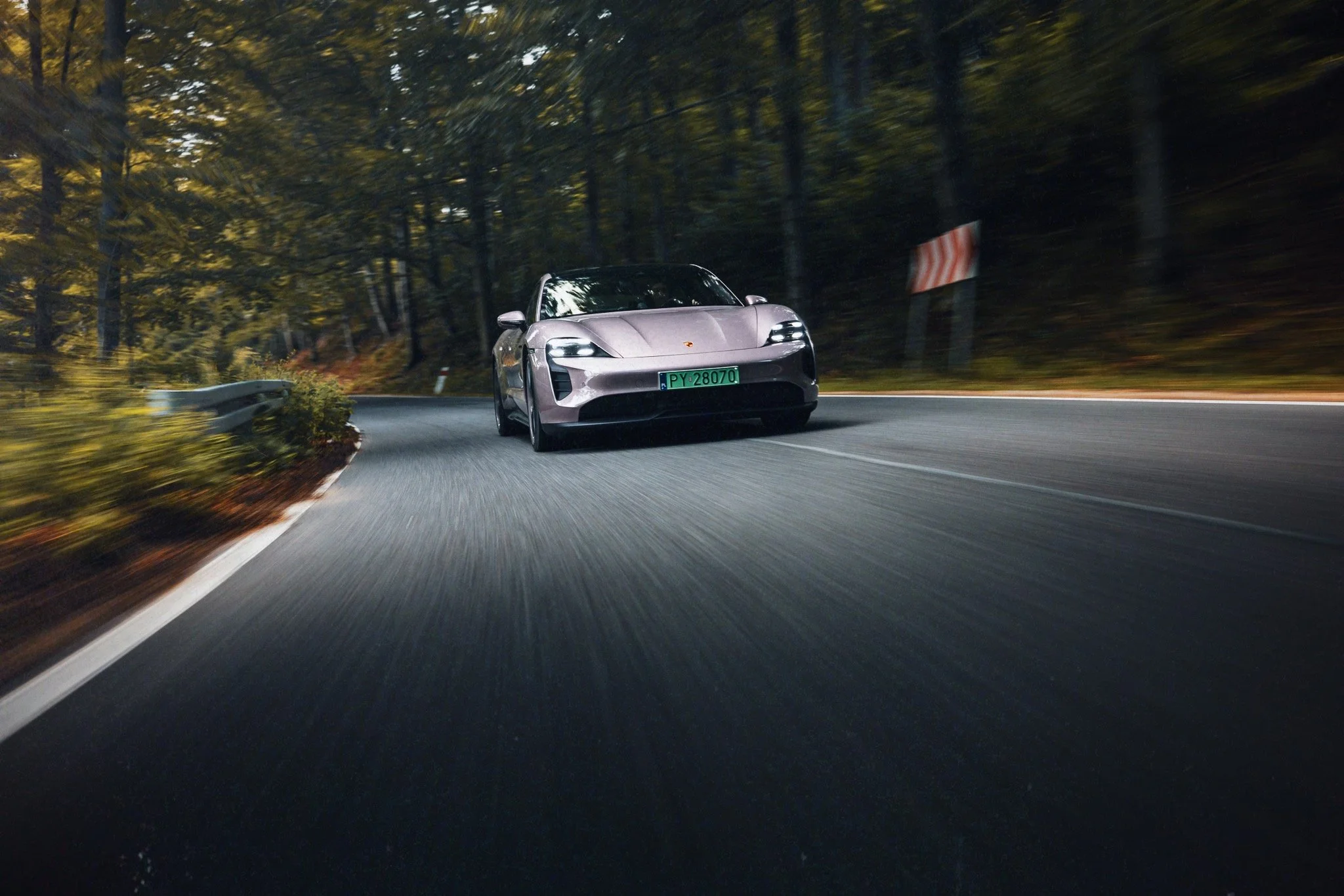 A silver sports car driving around a sharp bend on a winding road through a wooded forest at dusk.