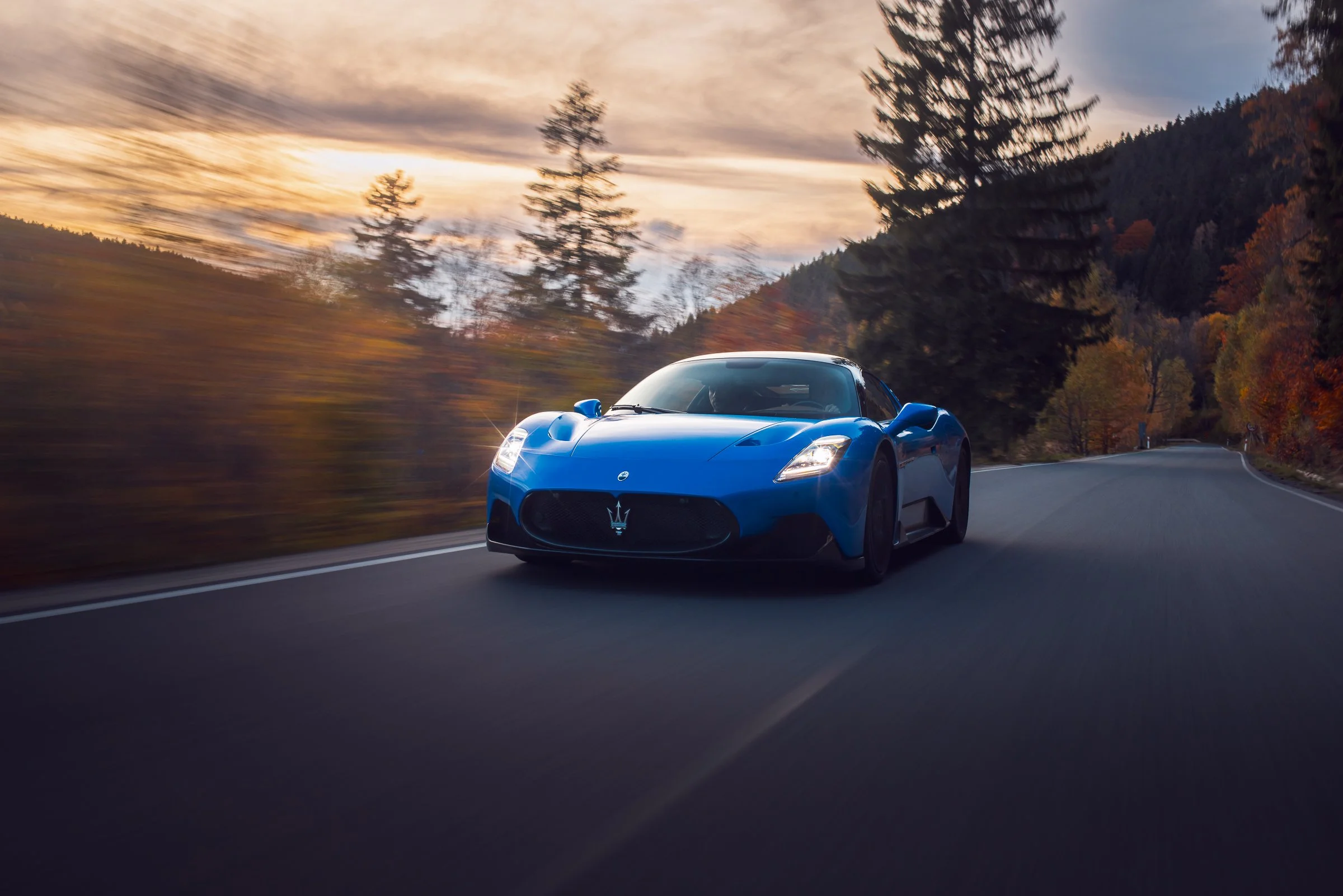 A blue sports car driving on a winding mountain road during sunset, surrounded by trees with autumn foliage.