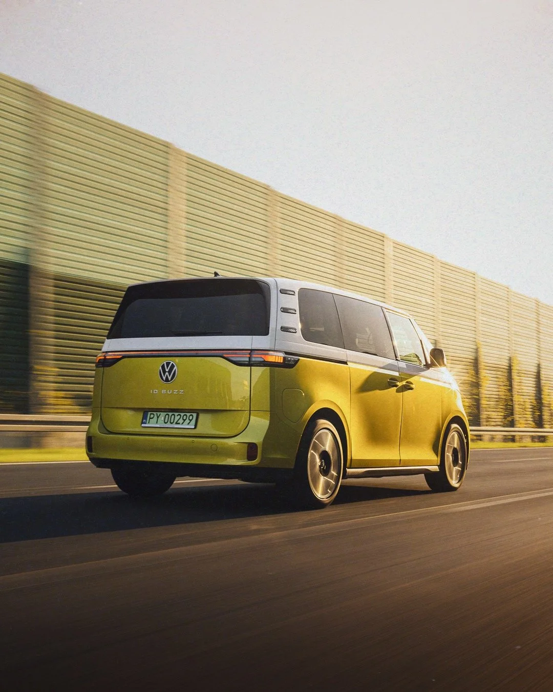 Yellow Volkswagen ID Buzz electric minivan driving on a highway with a sound barrier wall on the side.