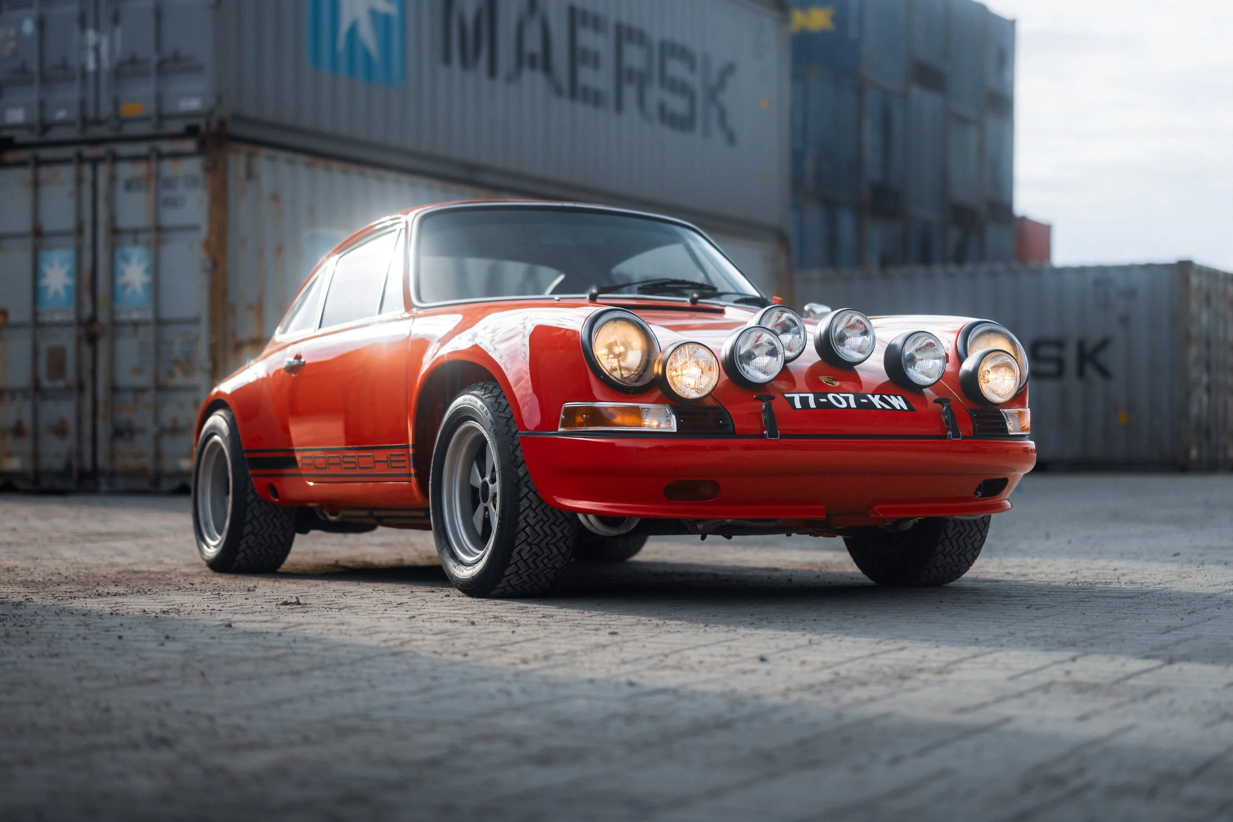 A vintage red Porsche sports car with multiple auxiliary lights on the front, parked on a concrete surface with shipping containers in the background.
