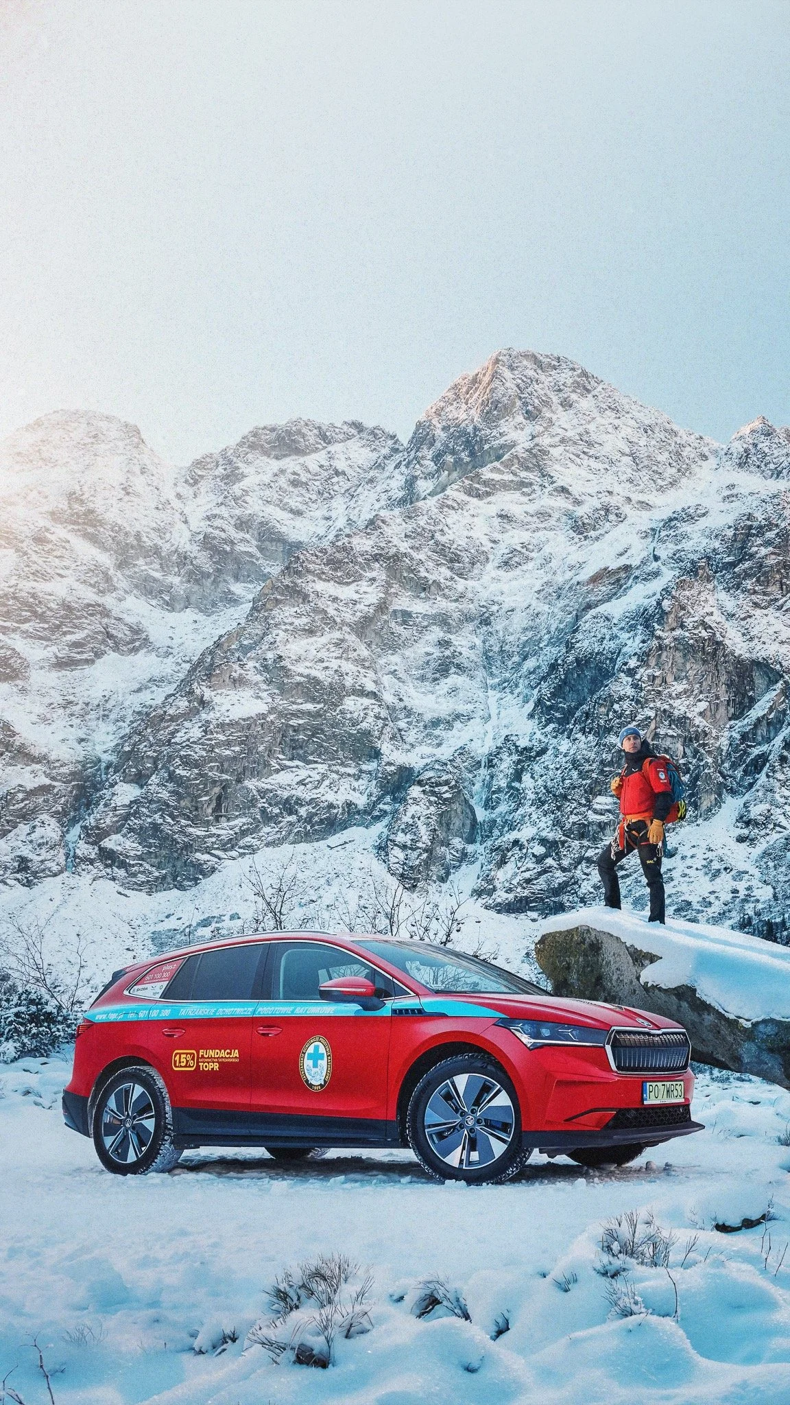 A red emergency vehicle parked in a snowy mountain landscape near a climber in winter gear, with tall snow-covered mountains in the background.