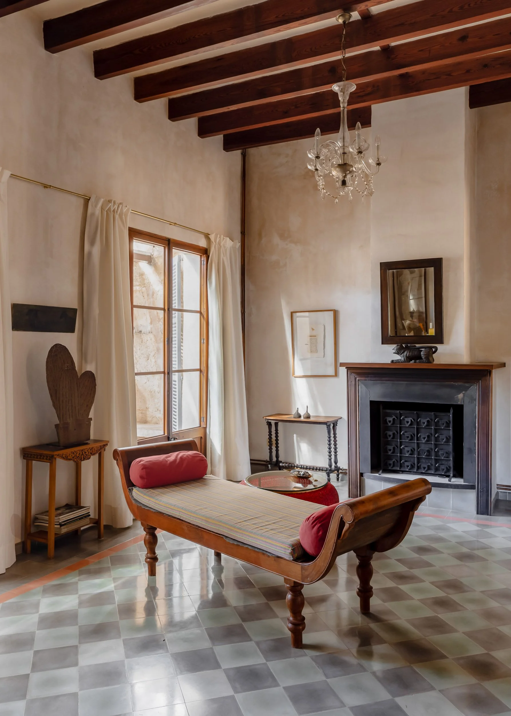 Sunlit Mediterranean interior with exposed wooden beams, a vintage chaise lounge with red bolsters, patterned tile floor and a classic fireplace beneath a crystal chandelier.