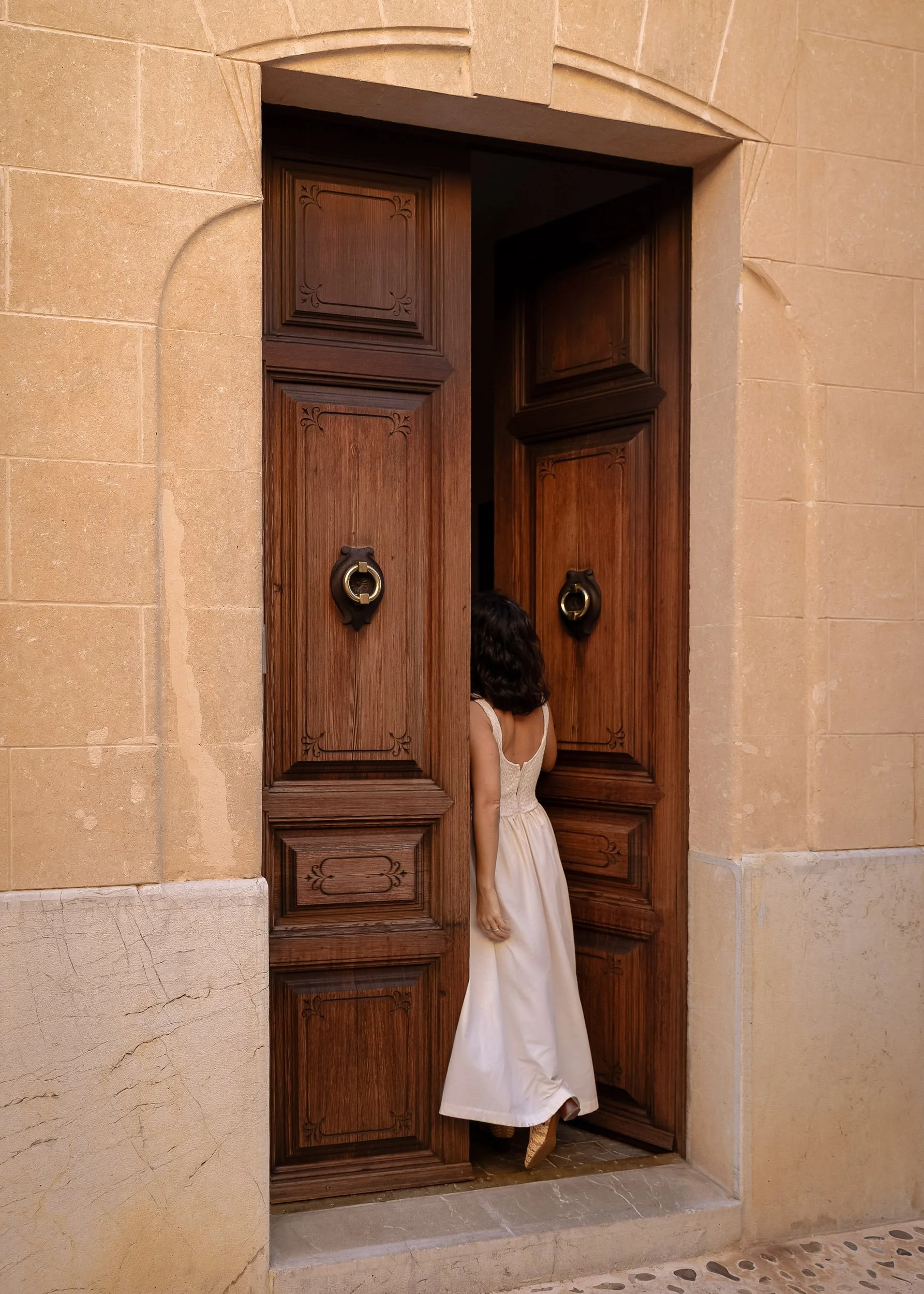 Woman in a white dress entering a large wooden door set in warm sandstone architecture, captured in soft natural light.