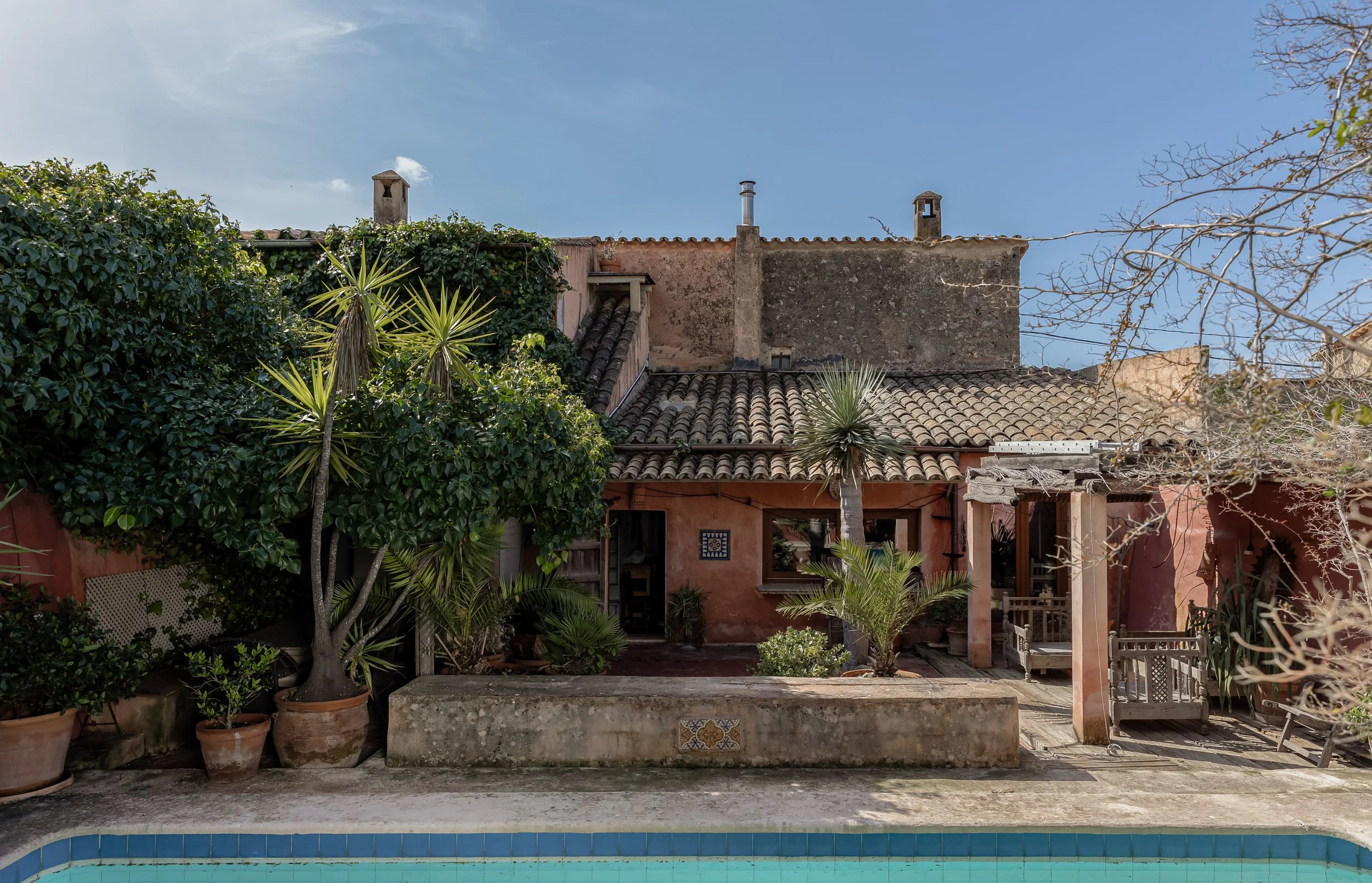 Wide angle architectural shot of a traditional Mallorcan stone house featuring a glimpse of a turquoise pool line and lush garden landscaping.