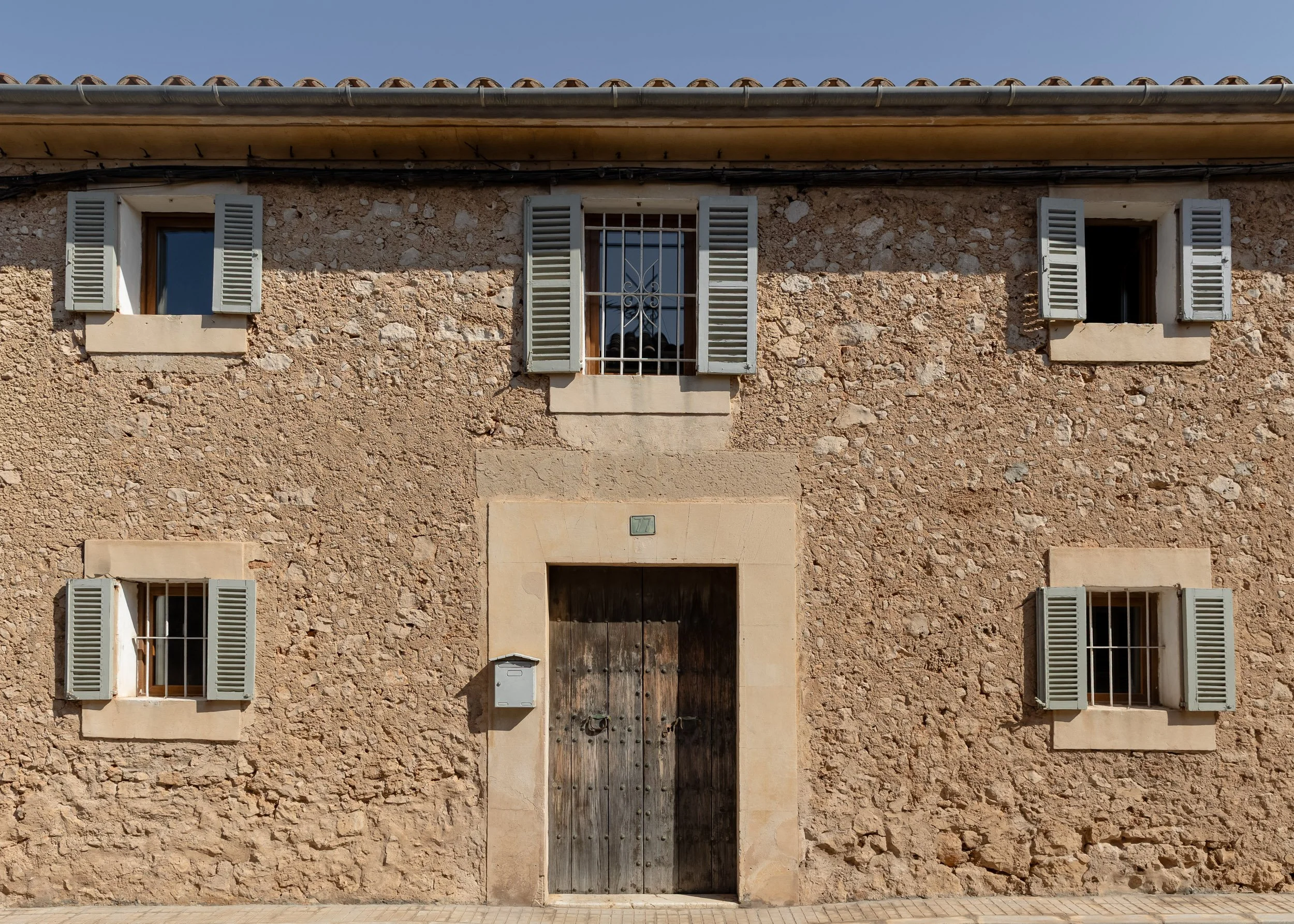 A rustic stone house with a wooden door, five windows with gray shutters, and a tiled roof under a clear blue sky.