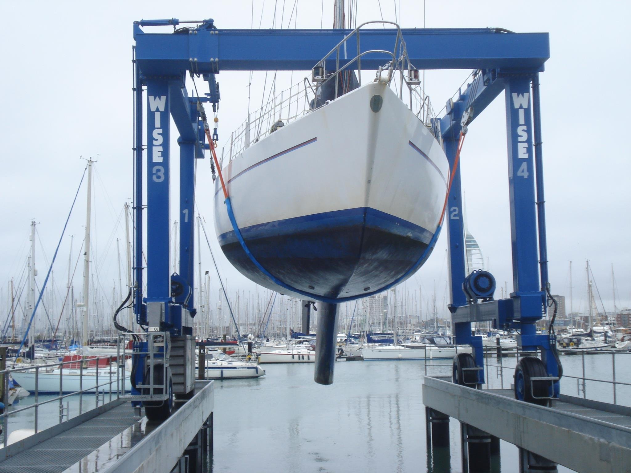 A boat hoist lifting a sailboat out of the water at a marina, with numerous other sailboats moored in the background.
