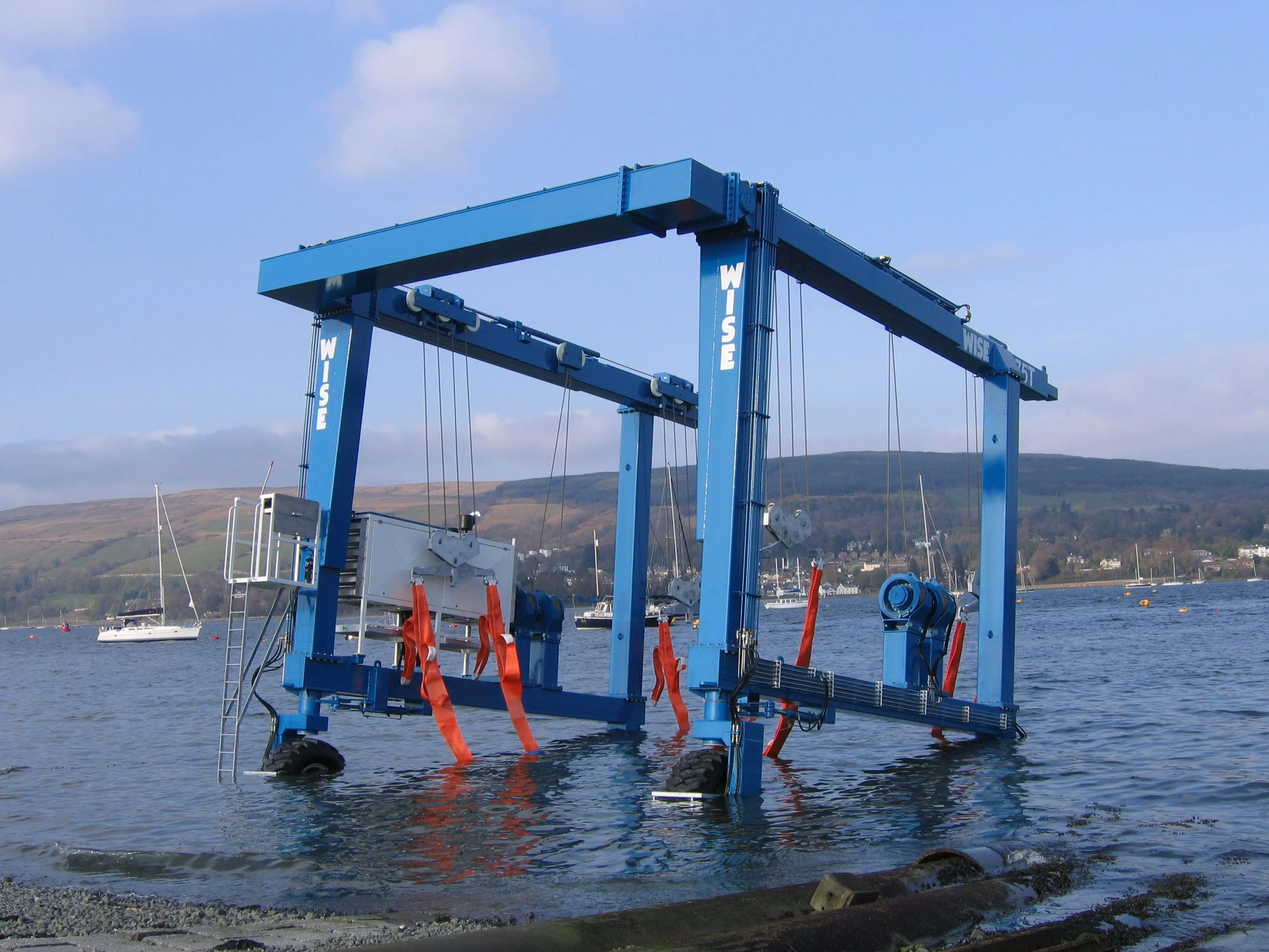 Large blue underwater lift device in a body of water with sailboats and hills in the background.
