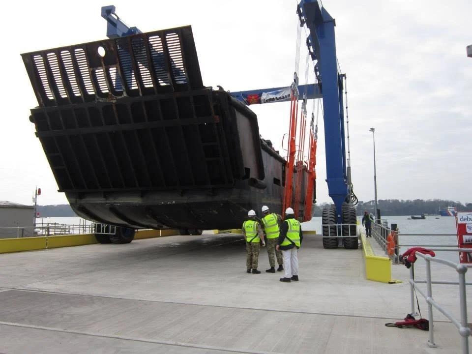 A large piece of equipment, possibly a dock or marine structure, being lifted by a blue crane held by four workers in safety vests and helmets at a dockyard by water.