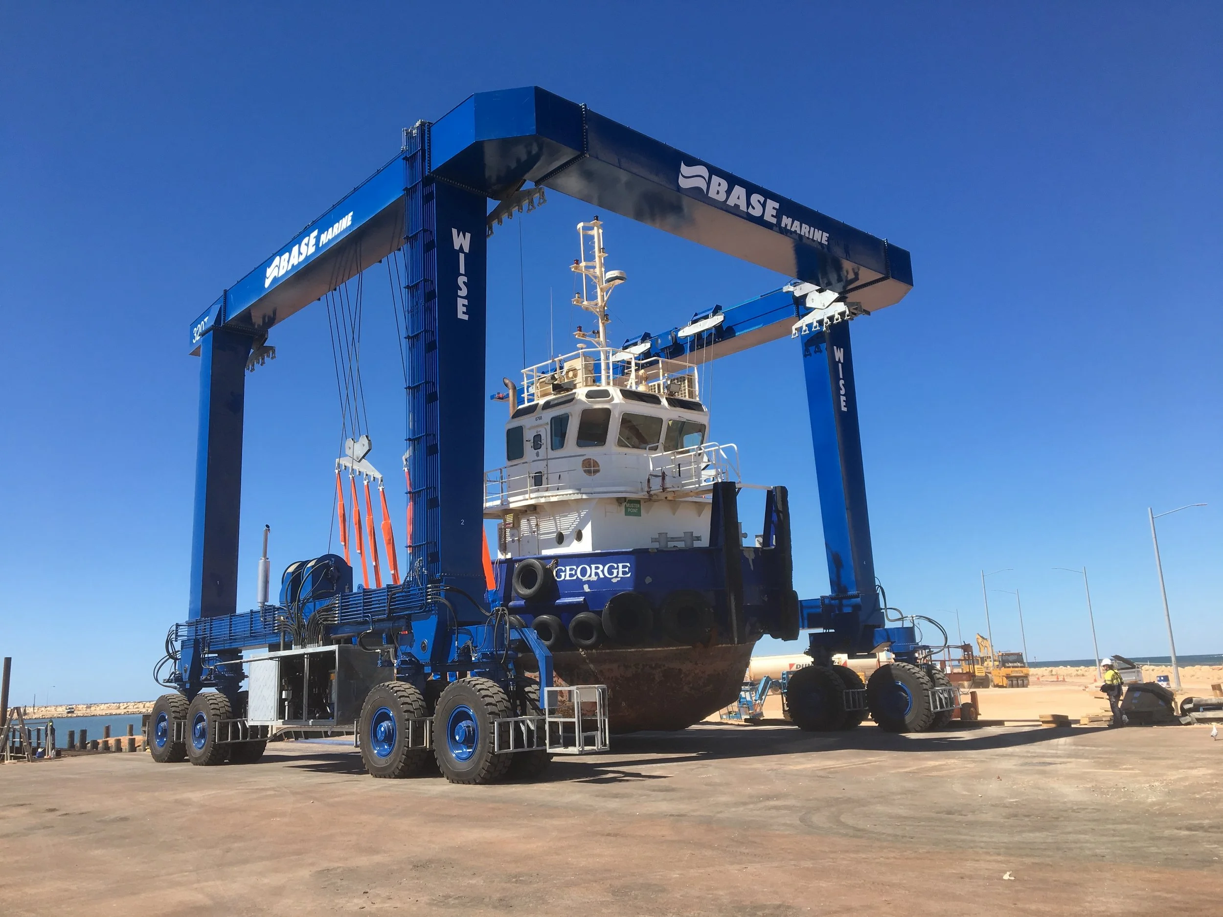 A large boat is suspended above the ground by a blue WISE Boat Hoist at a boat yard in Australia