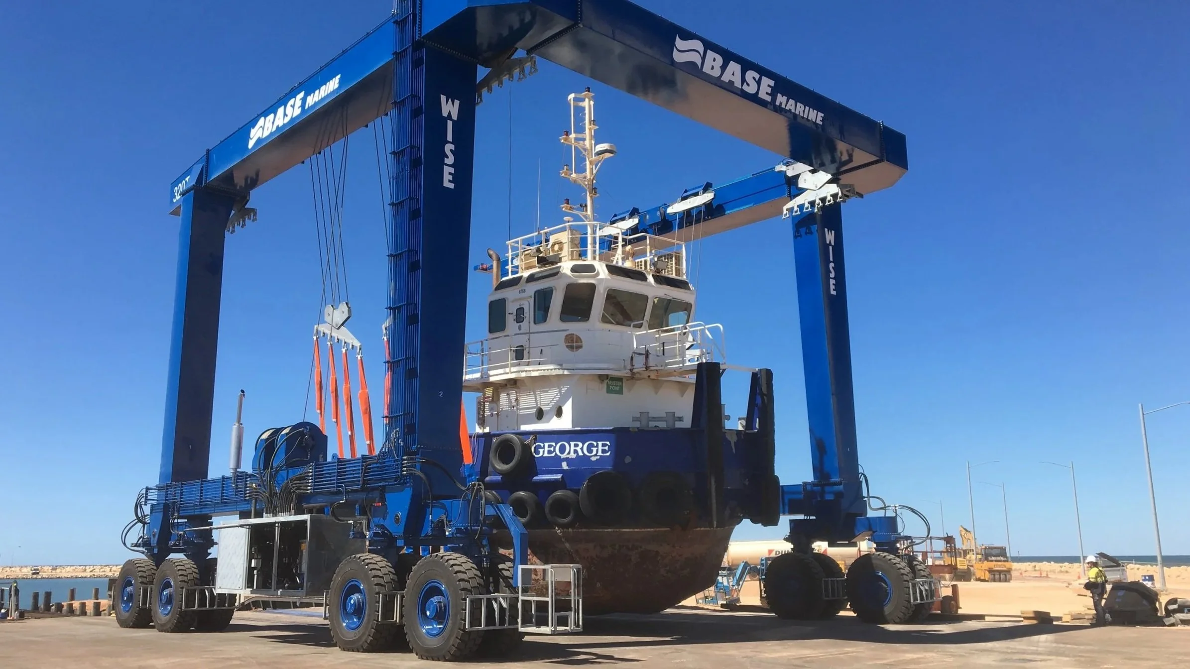 A large boat is suspended above the ground by a blue WISE Boat Hoist at a boat yard in Australia