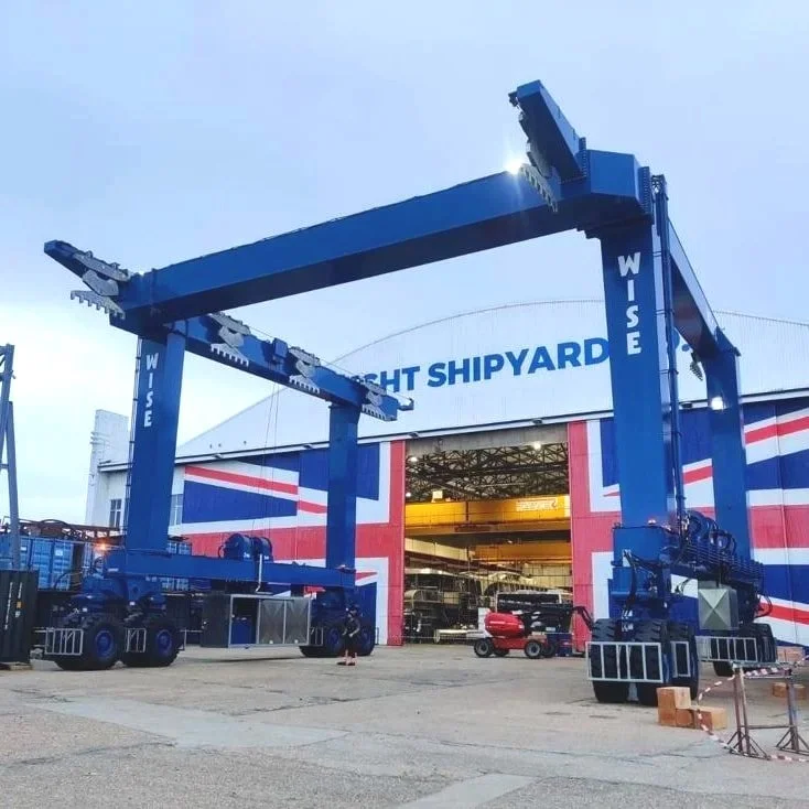 Large blue gantry cranes labeled 'Wise' in front of a shipyard building with 'Night Shipyard' sign, used for lifting ships or heavy materials.