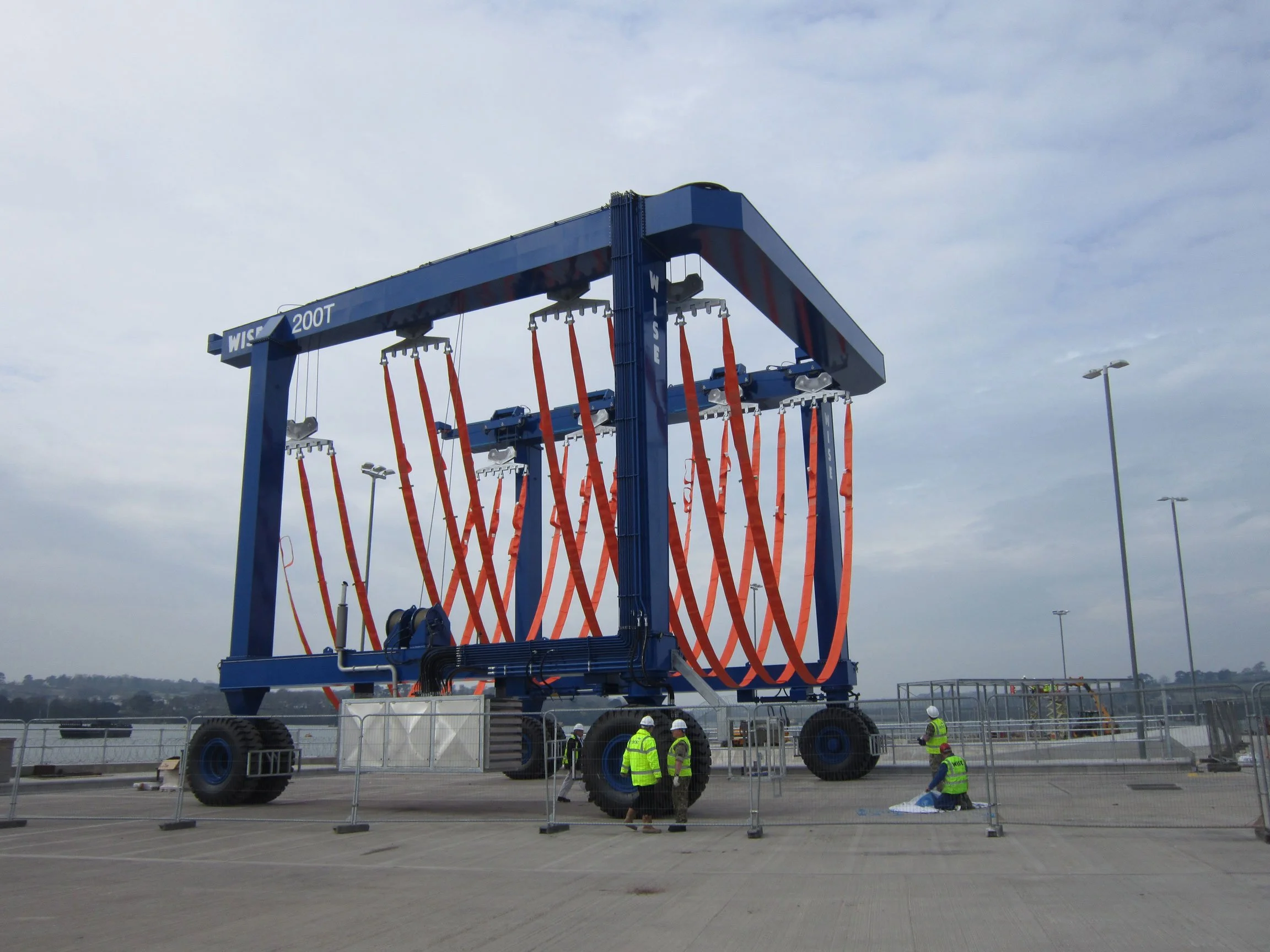 Large blue and red wave energy testing machine on a flat surface, surrounded by construction barriers, with workers in safety vests and helmets nearby, and a cloudy sky overhead.